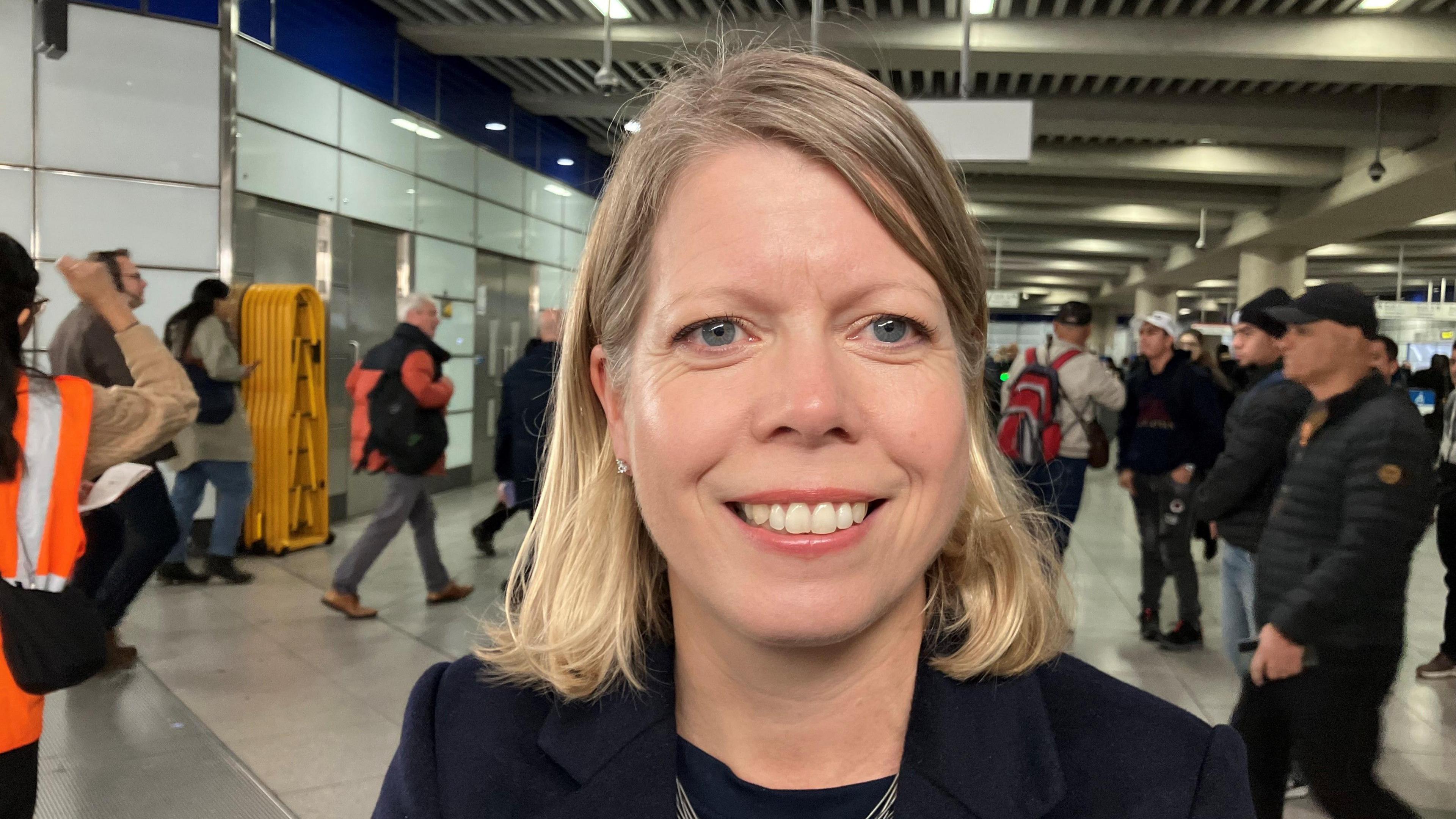 A blonde woman in navy business clothes smiles at a railway station.