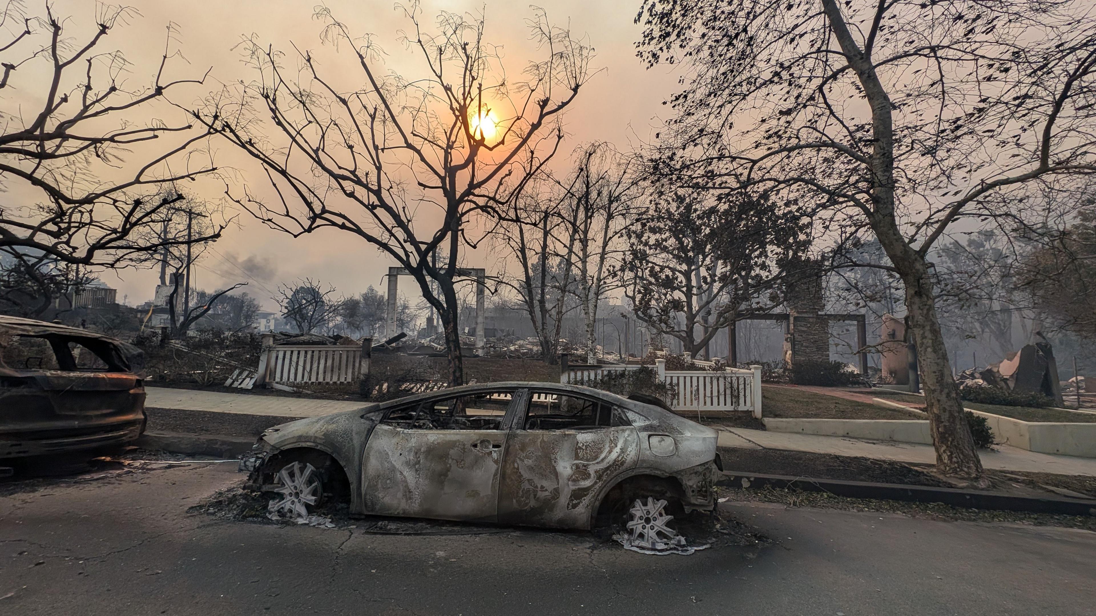 A burned out car surrounded by burnt buildings as the sun shines through smokey clouds