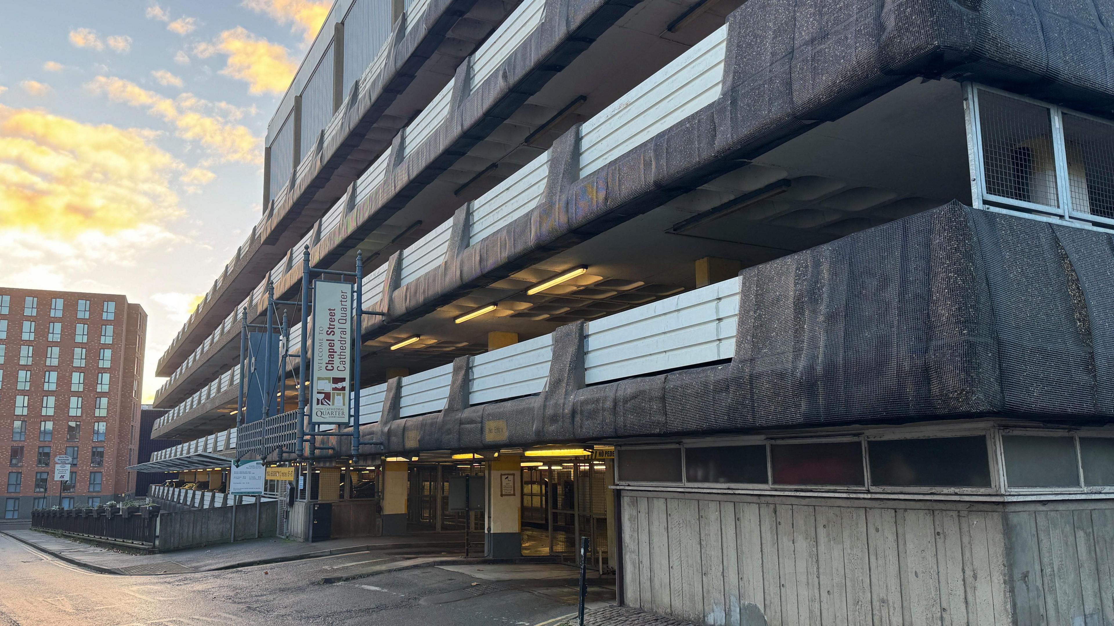 Exterior of a concrete multi-storey car park, five floors are visible. A hanging sign says "Welcome to Chapel Street Cathedral Quarter"