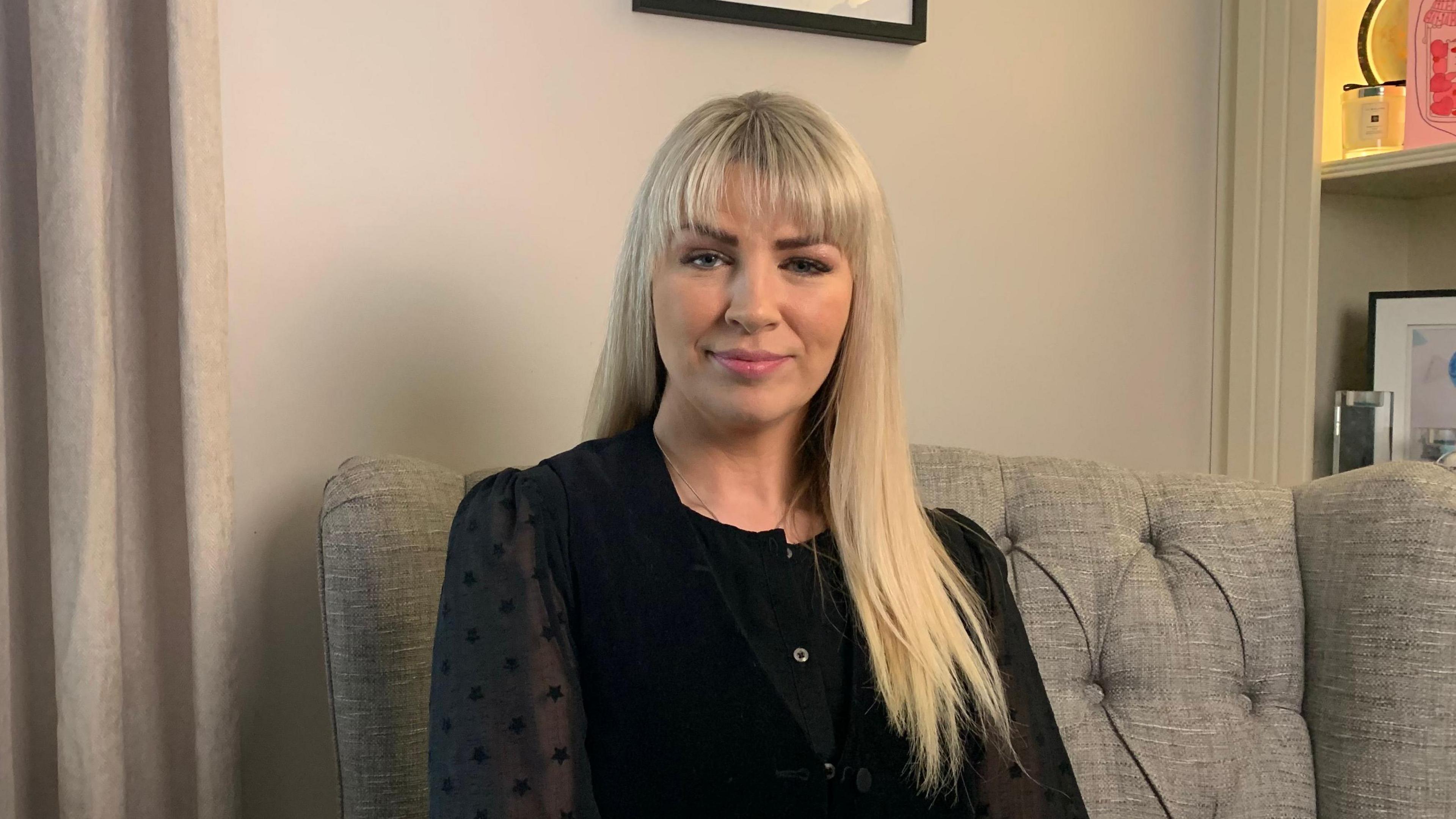 Marie is sitting in a grey armchair, looking directly at the camera with a slight smile. She is wearing a black long sleeved top and has long blonde hair with a fringe. 