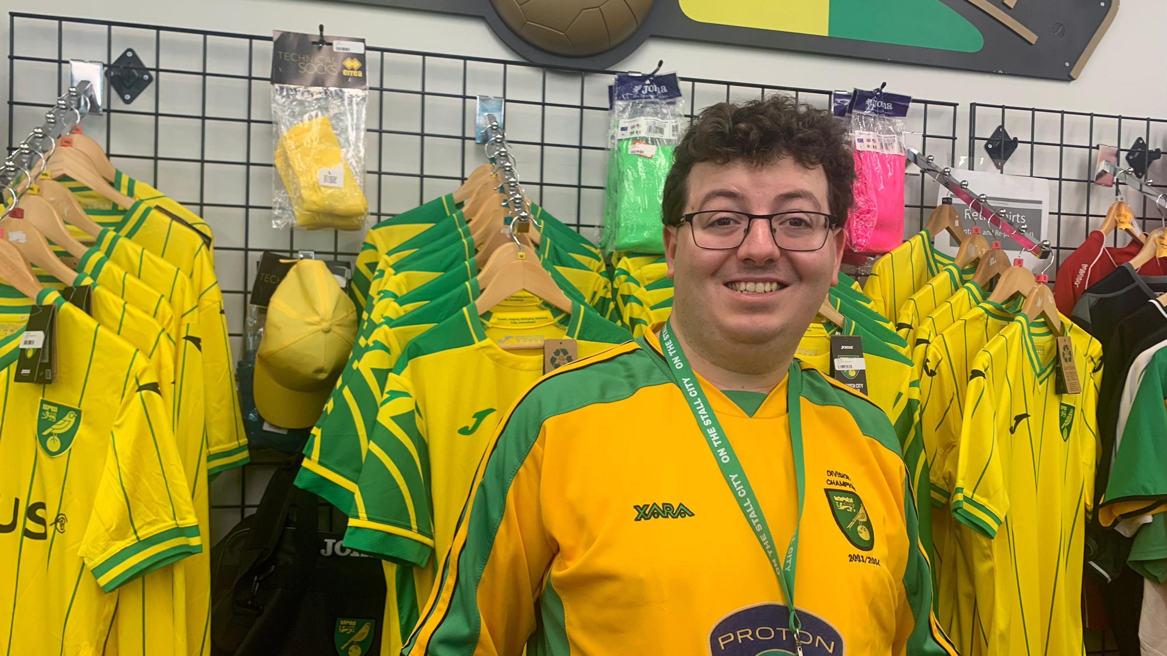 A man, who is smiling, and is wearing a green lanyard and a yellow and green football shirt. There are racks of the same shirts behind him.