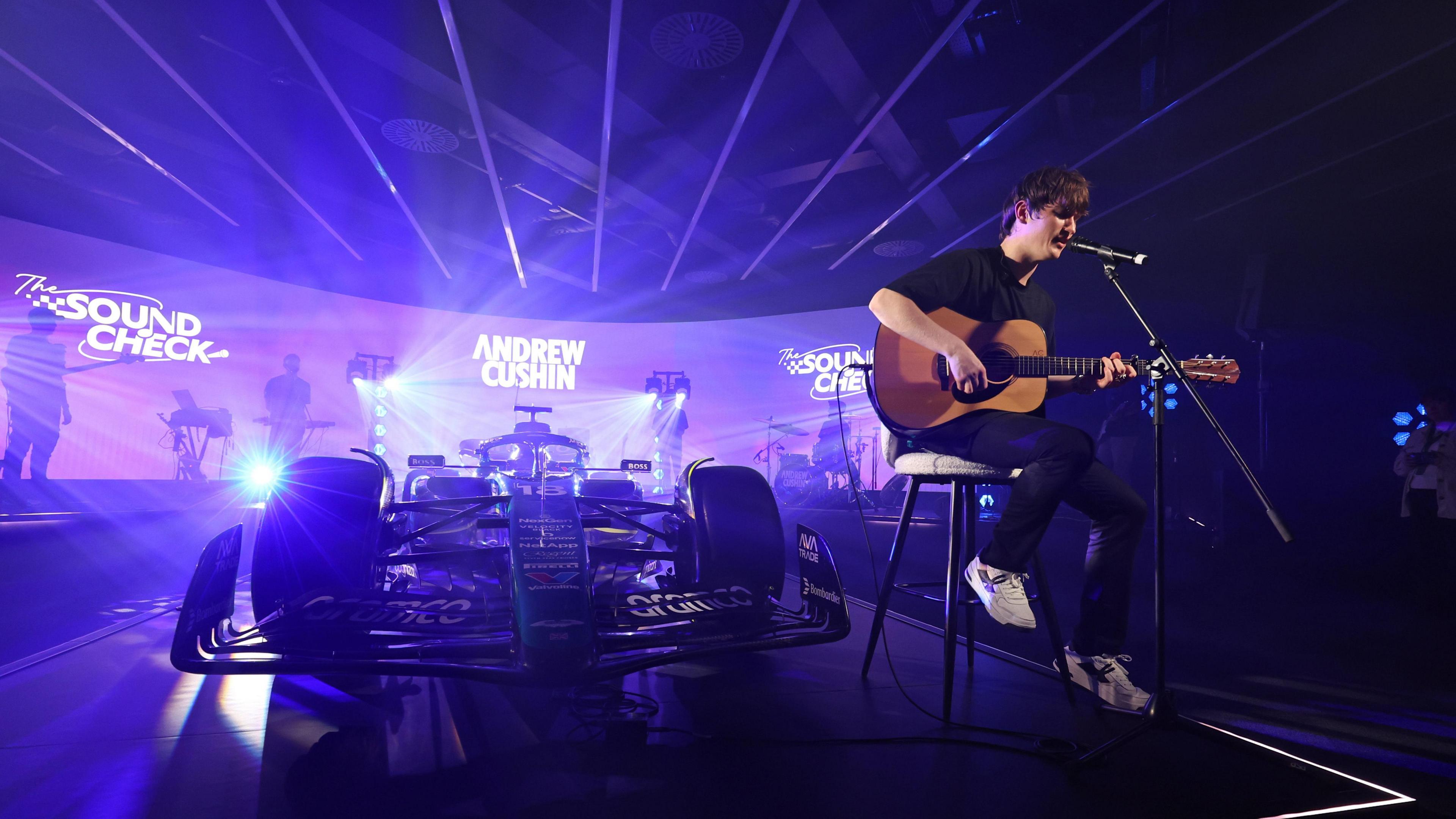 Andrew Cushin sitting on a stool with an acoustic guitar and mic. Behind him is an Aston Martin F1 car bathed in violet light.