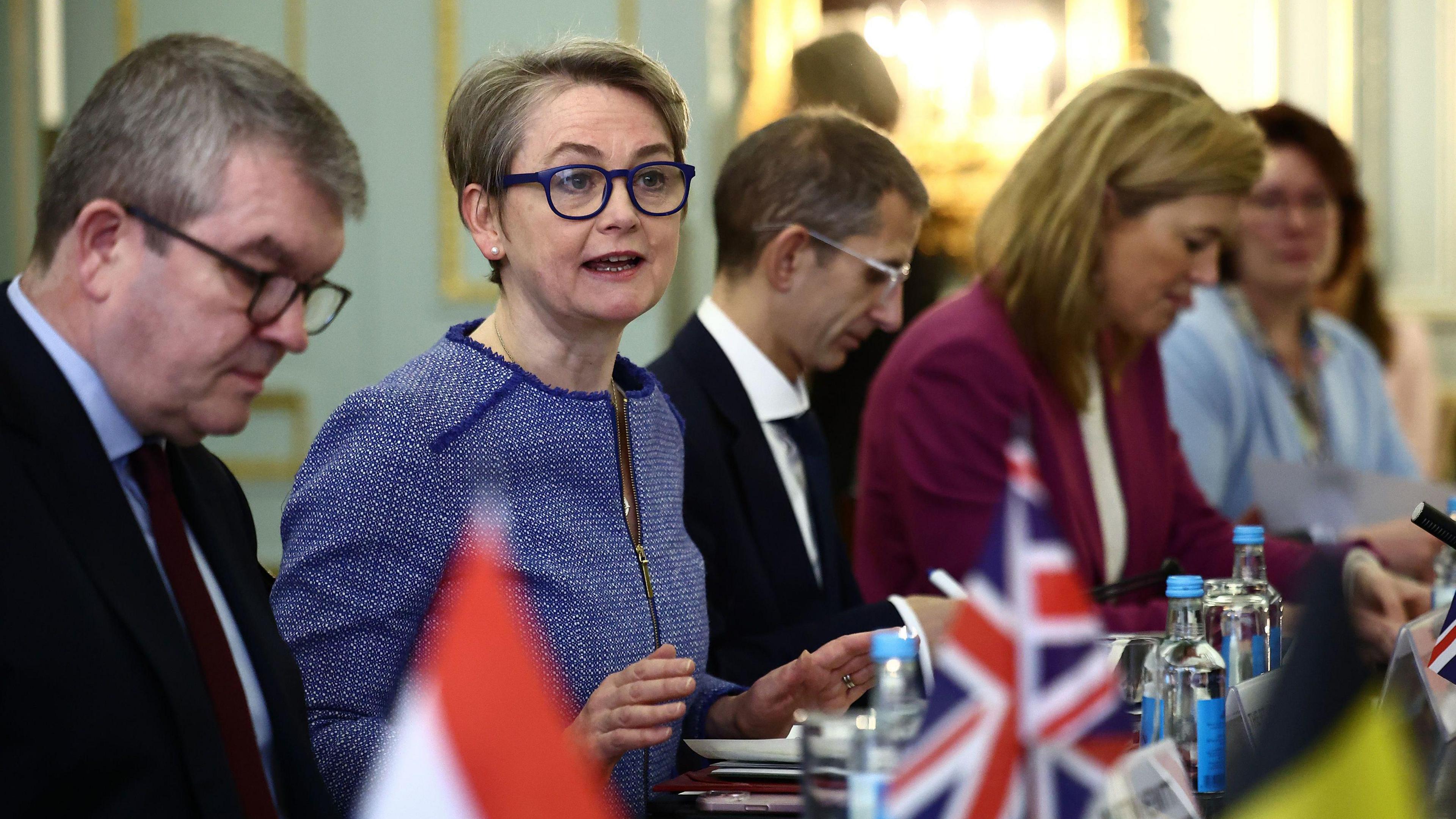 Yvette Cooper speaking around table. There are four other people seen sat on her side of the table, with small flags sitting on top.