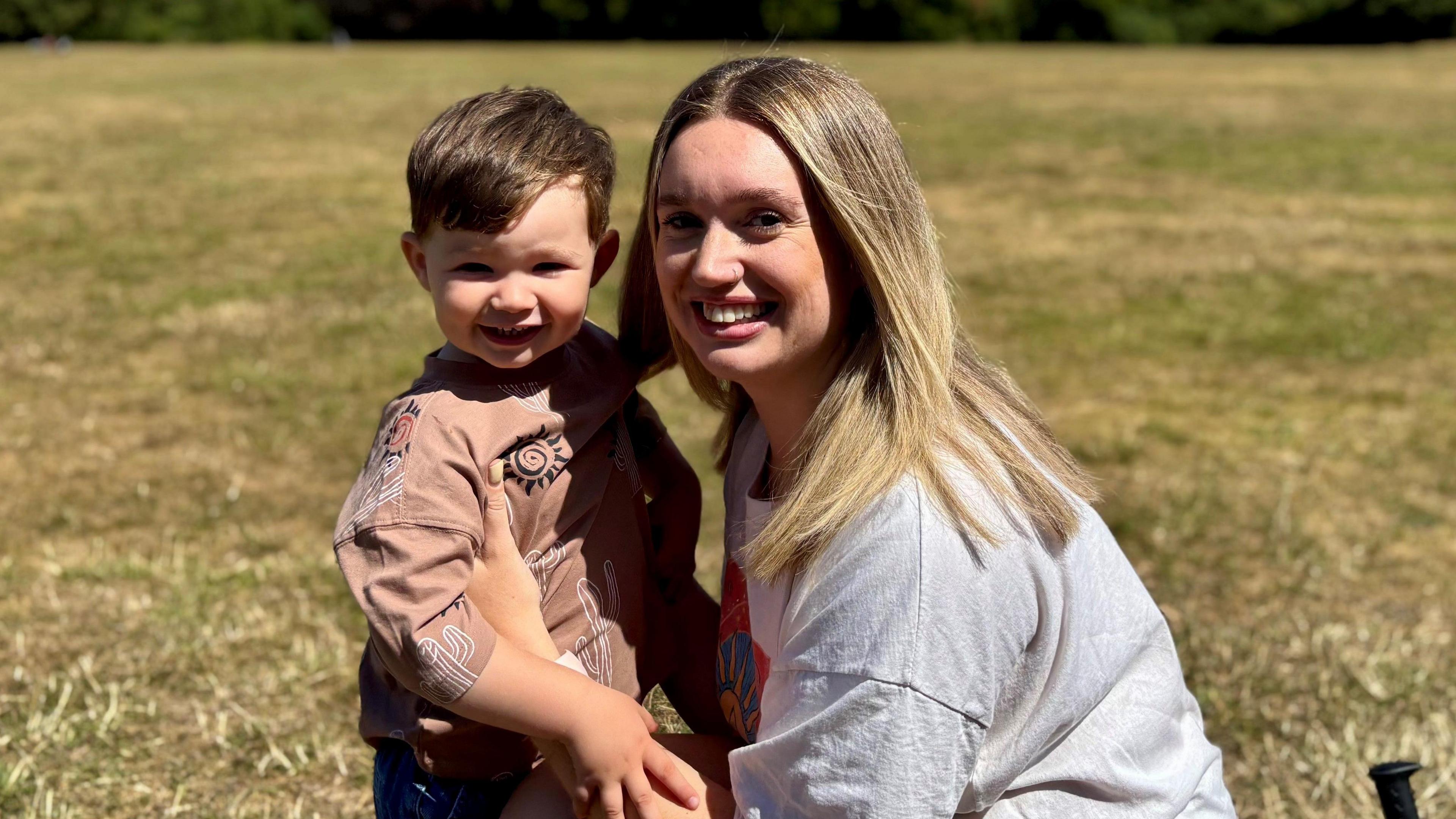 Karis in. a grey T-shirt, holding two-year-old Noah, standing on a lawn. Both are smiling.
