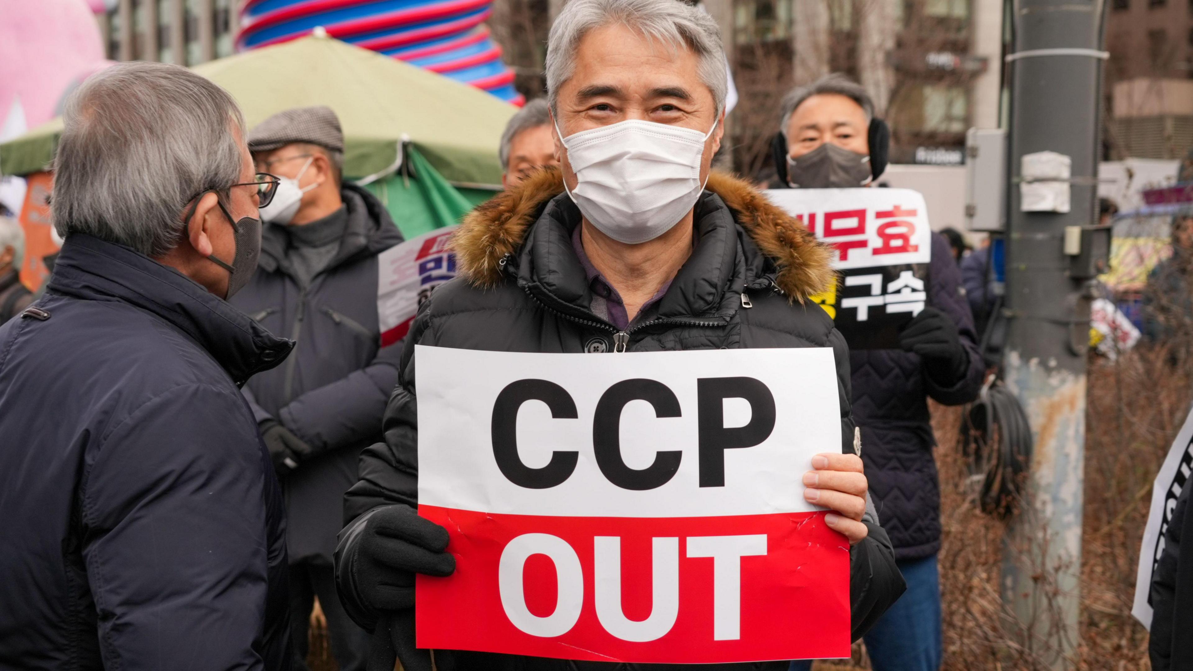 A man wearing a disposable face mask holds a sign that reads "CCP OUT"