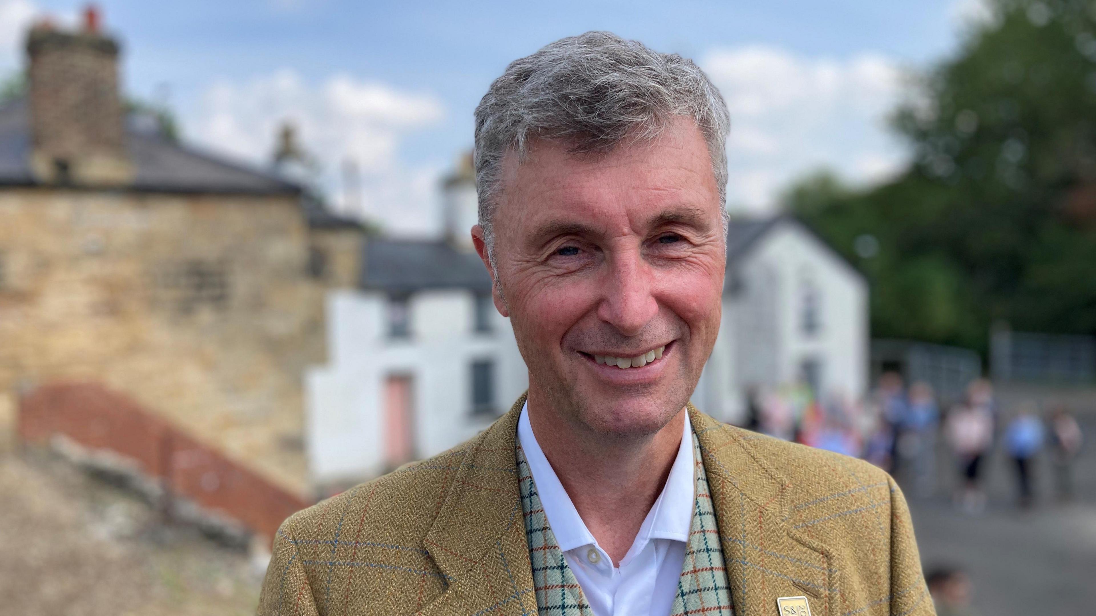 Niall Hammond with grey hair, wearing a mustard tweed blazer and white shirt, smiles at the camera.