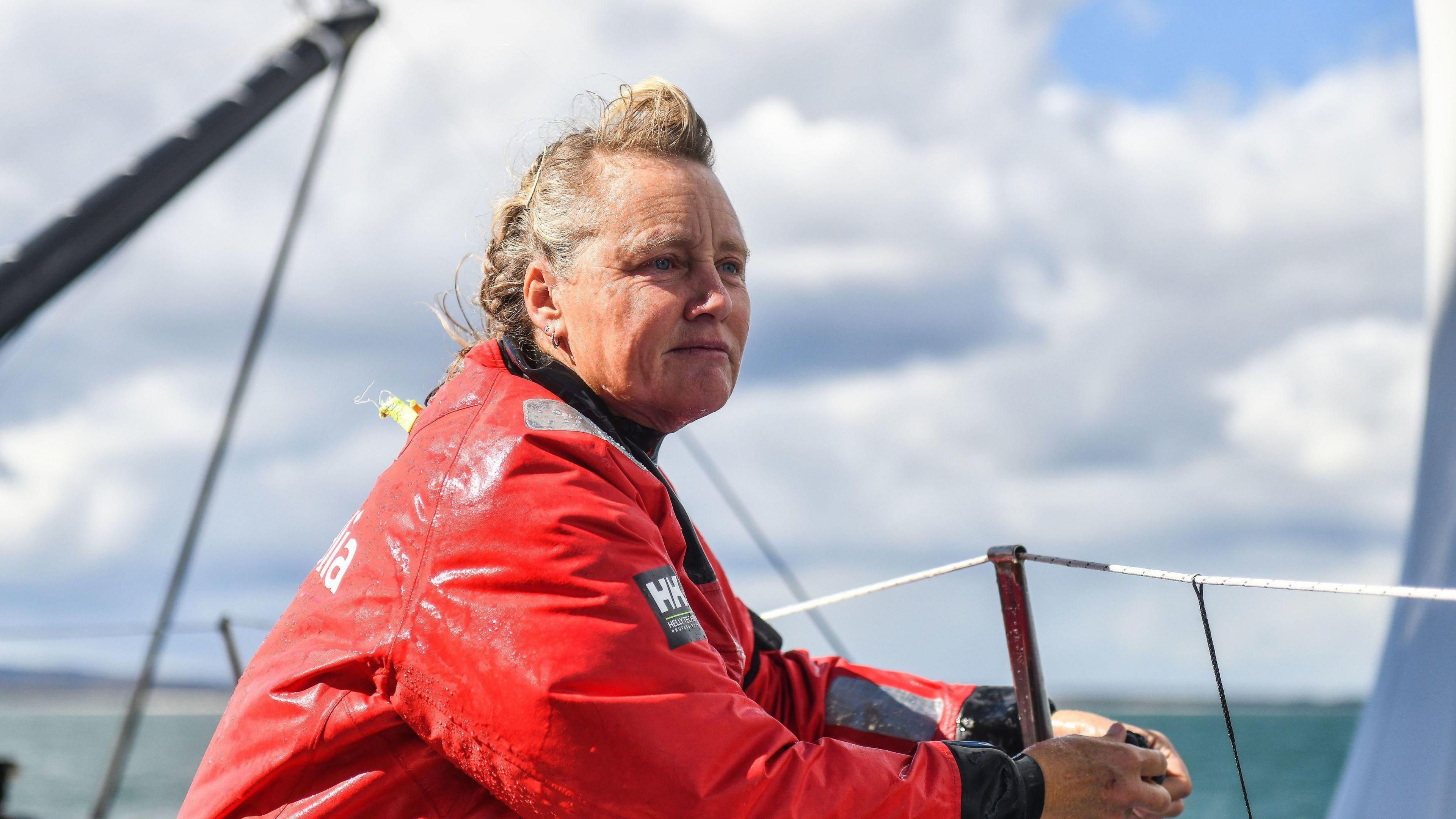 Pip Hare looks over the edge of her yacht toward the sea as she fastens a rope around a metal pole. Her blonde and grey hair is windswept and she is wearing a large red waterproof coat