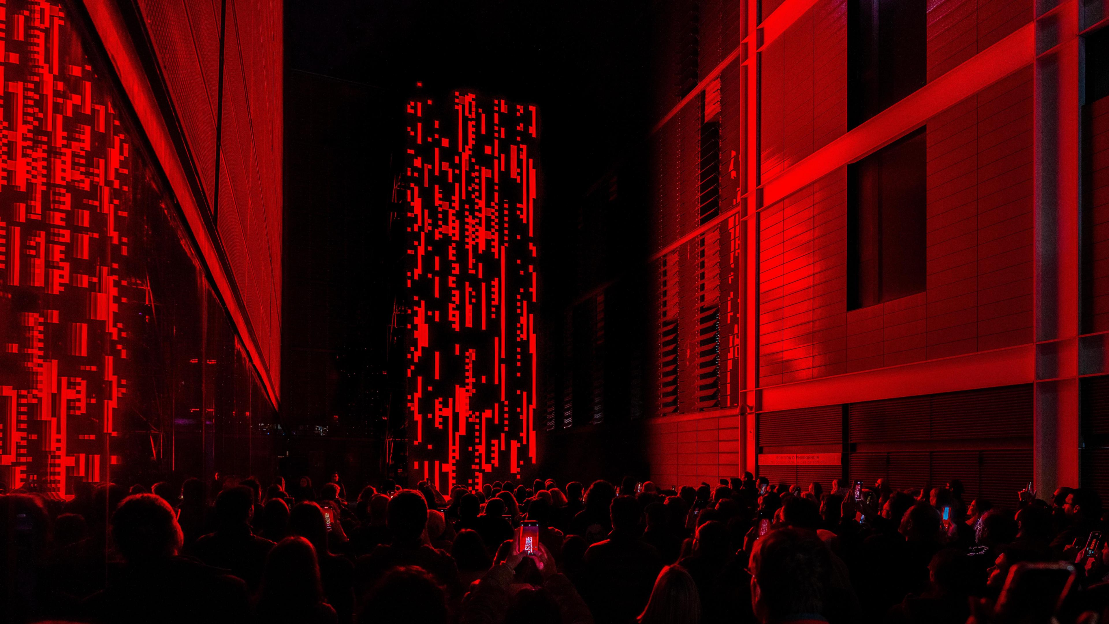 A large rectangular statue stands in between two buildings. It is black with red digital lines and shapes, and omits a red glow on the surrounding buildings. Silhouettes of people watching and taking photos are at the bottom of the picture.