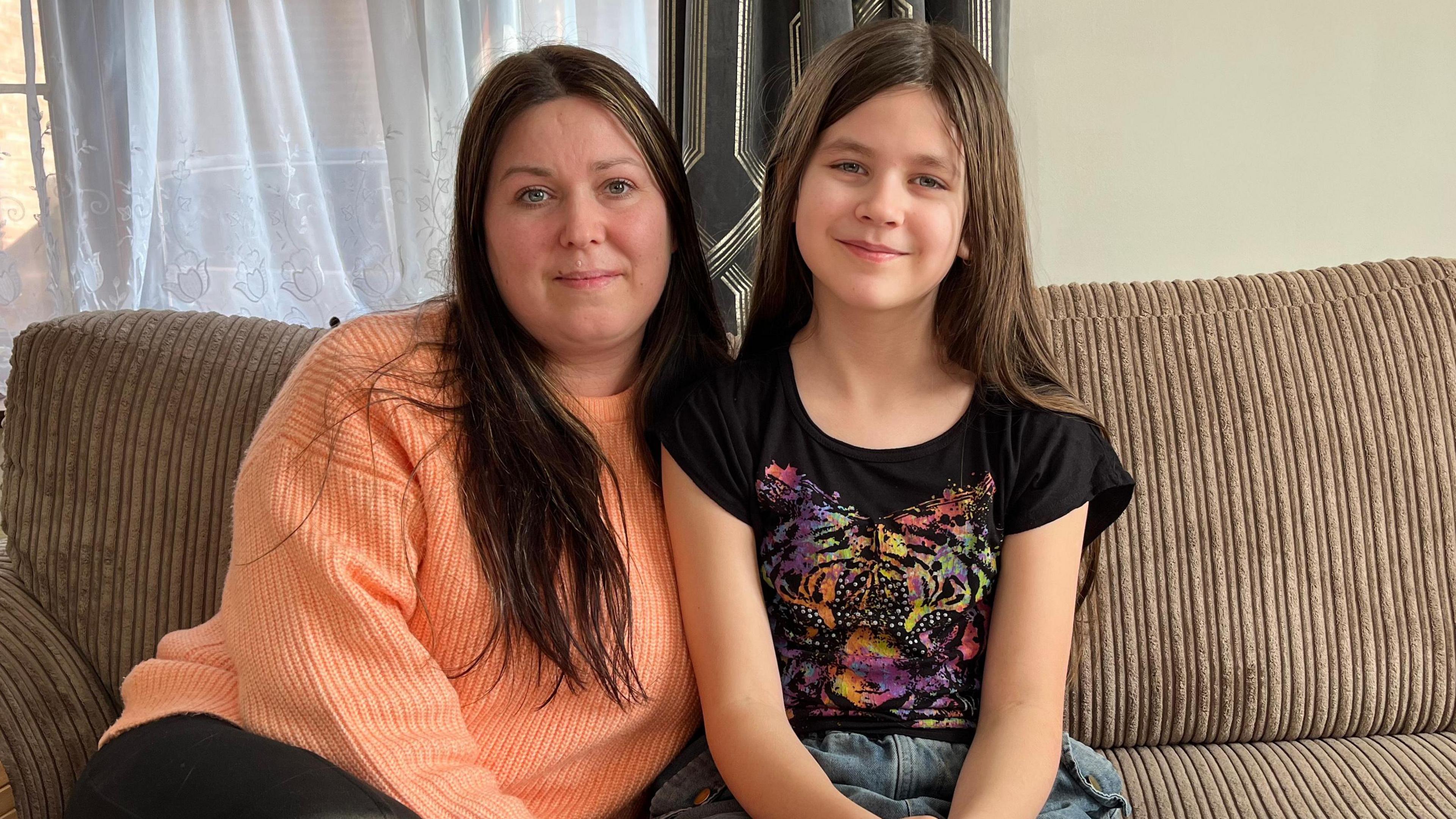 Svetlana wears an orange jumper and has long brown hair. Her 10 year old daughter Viktoria sits to her right. She is wearing a black t shirt with a multi coloured print of a tiger on it. She also has long brown hair. 