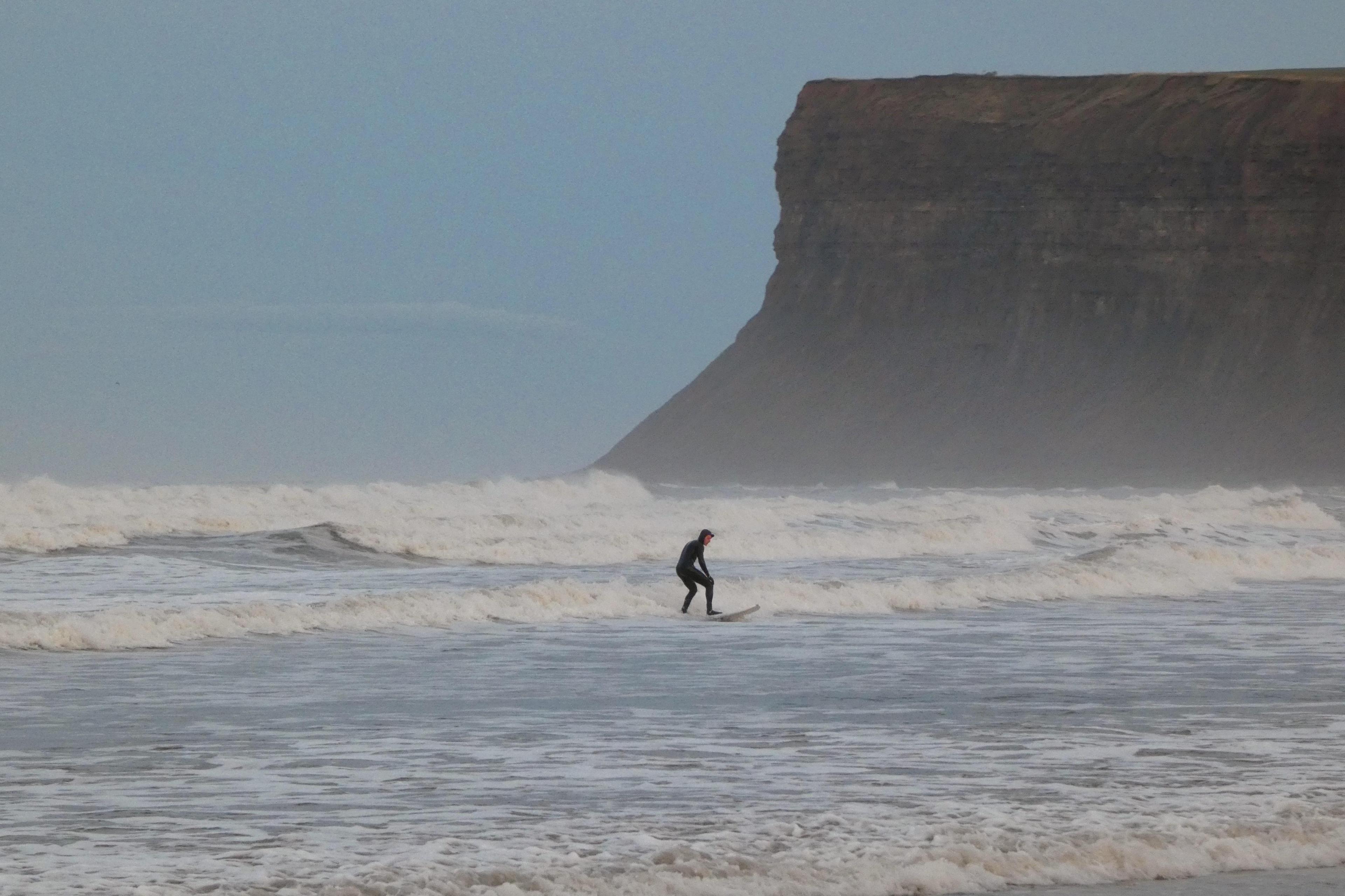 A surfer, in a full body-suit, rides a wave in front of a cliff.