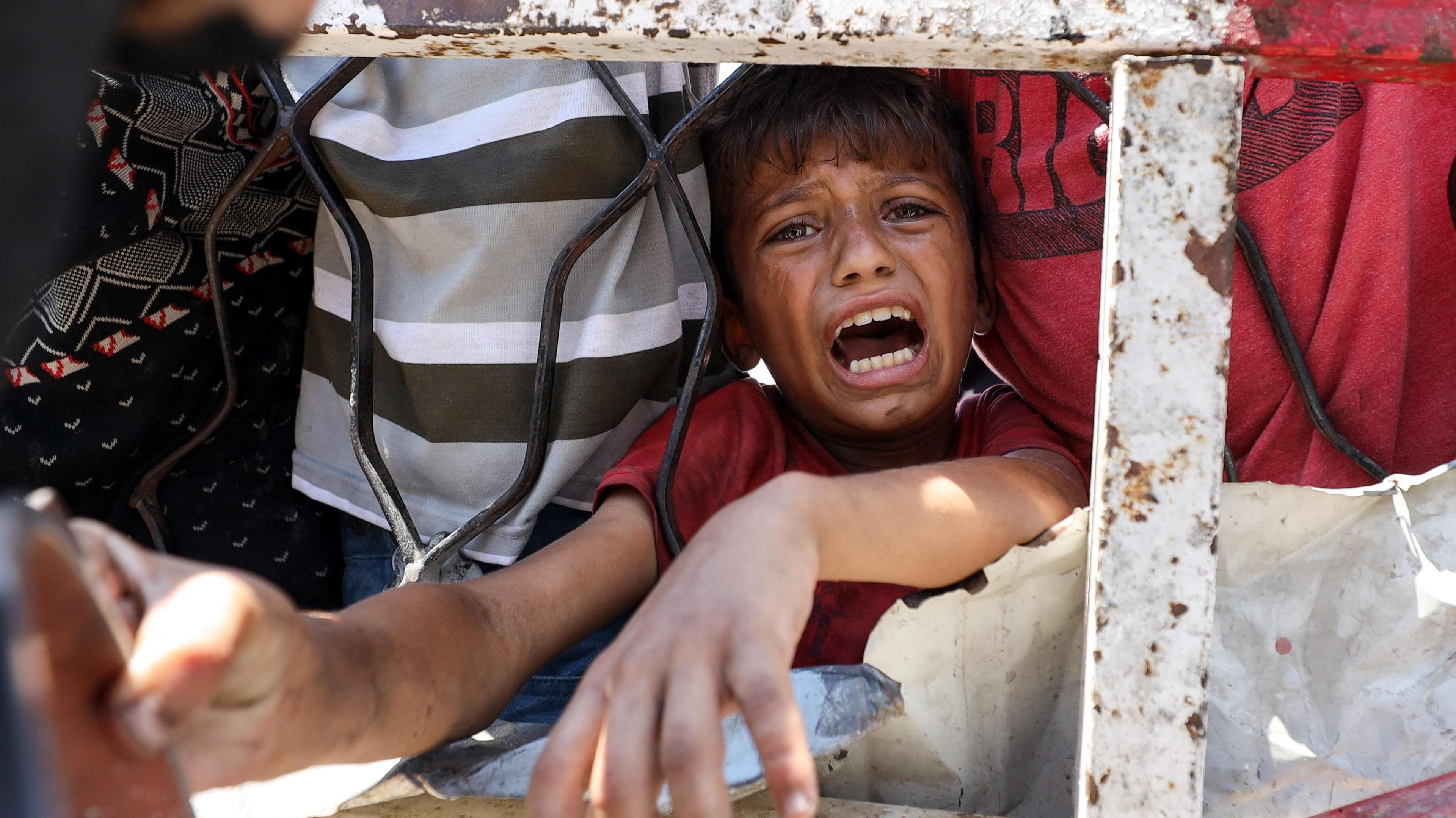A little boy cries as he waits for food at a charity kitchen, his arms pushed through a railing, clutching a pot, towards the camera.