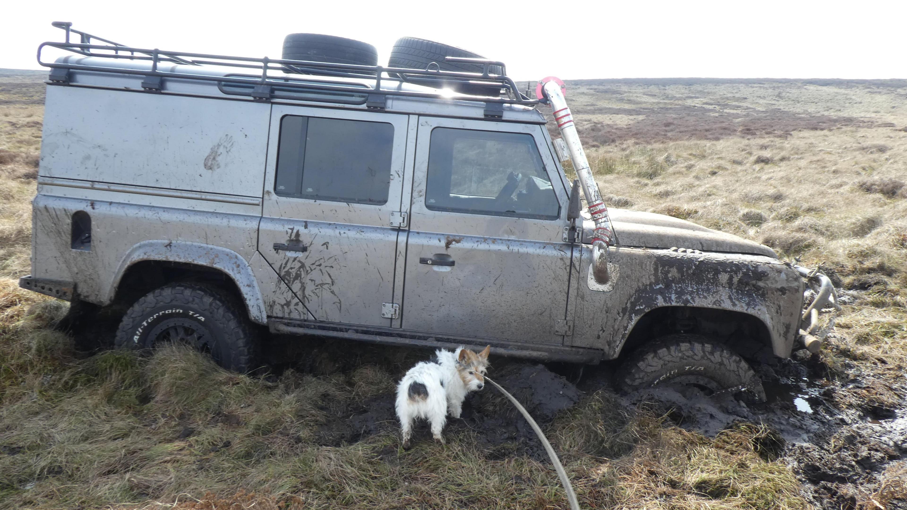 Picture shows a grey-silver coloured jeep stuck in mud on Ilkley Moor. Its wheels are almost completely submerged in the mud. A small dog stands in front of the scene and looks back.