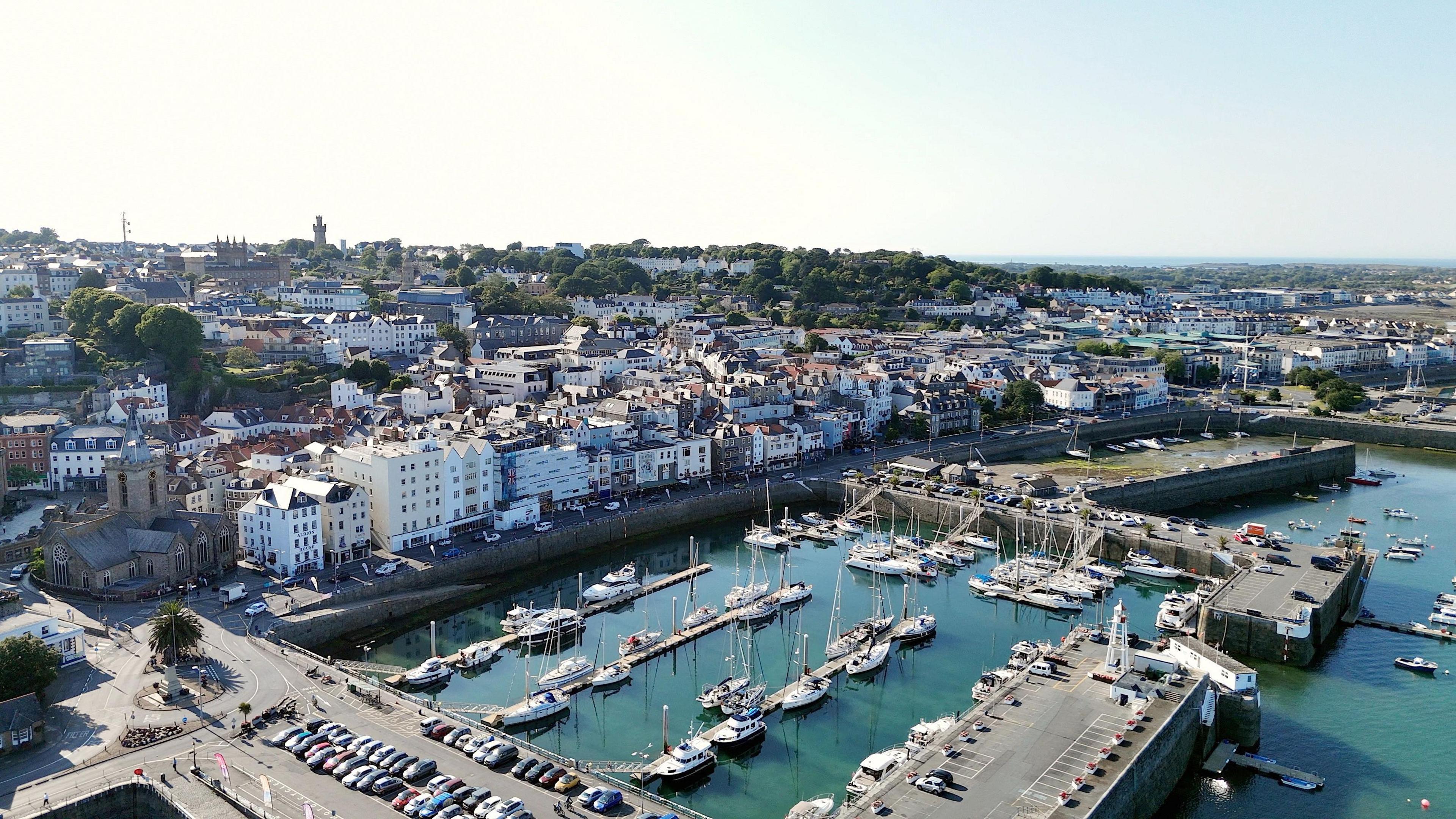 Aerial view of a harbour. Boats are moored in the water. Cars are parked in a car park next to the harbour. Properties are built up next to the water. Properties can be seen in the distance.