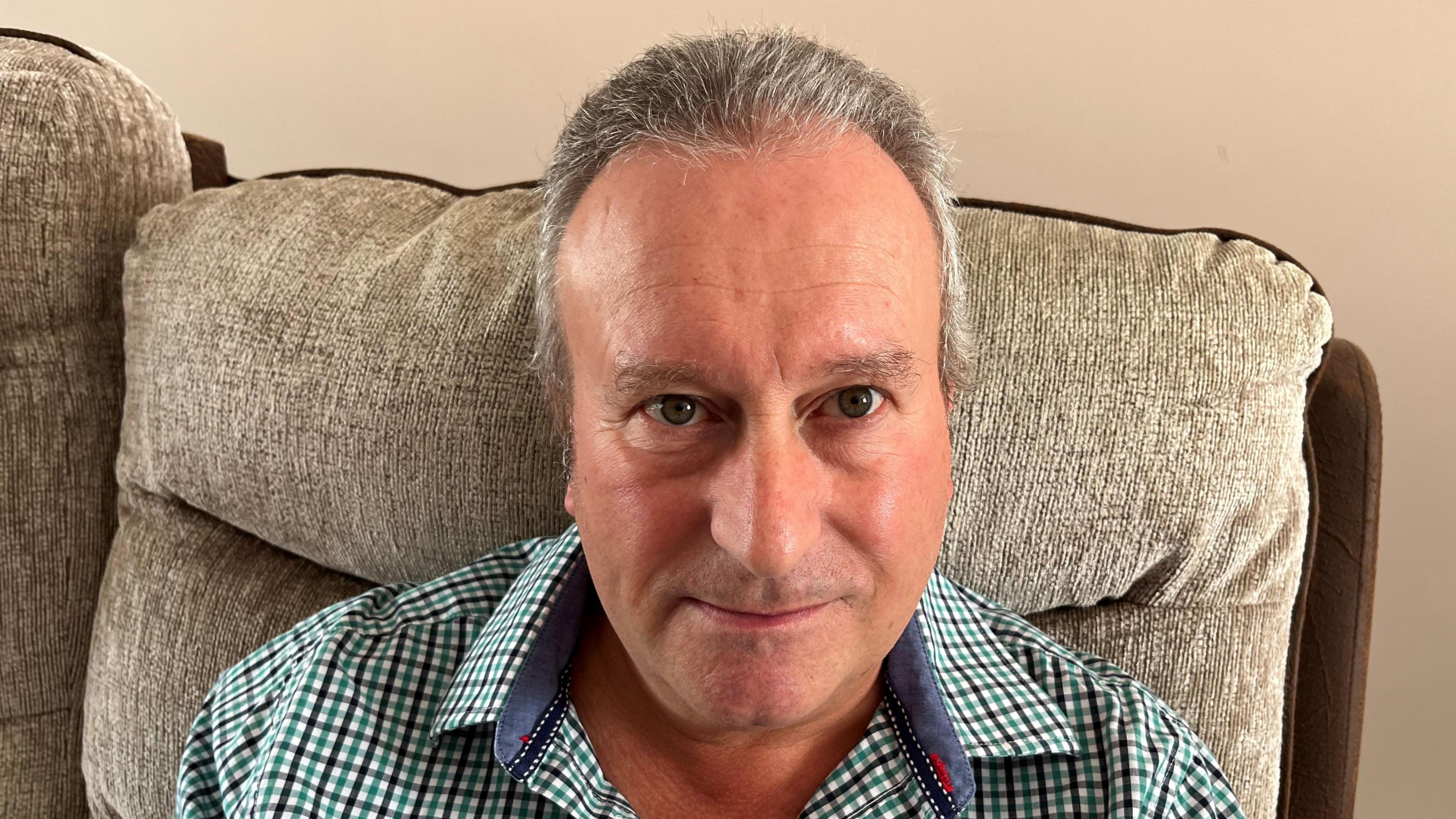 Darren Miles has short grey hair and a green checked shirt on with blue inside the collar. He is sitting in a cream coloured arm chair with a beige living room wall behind.