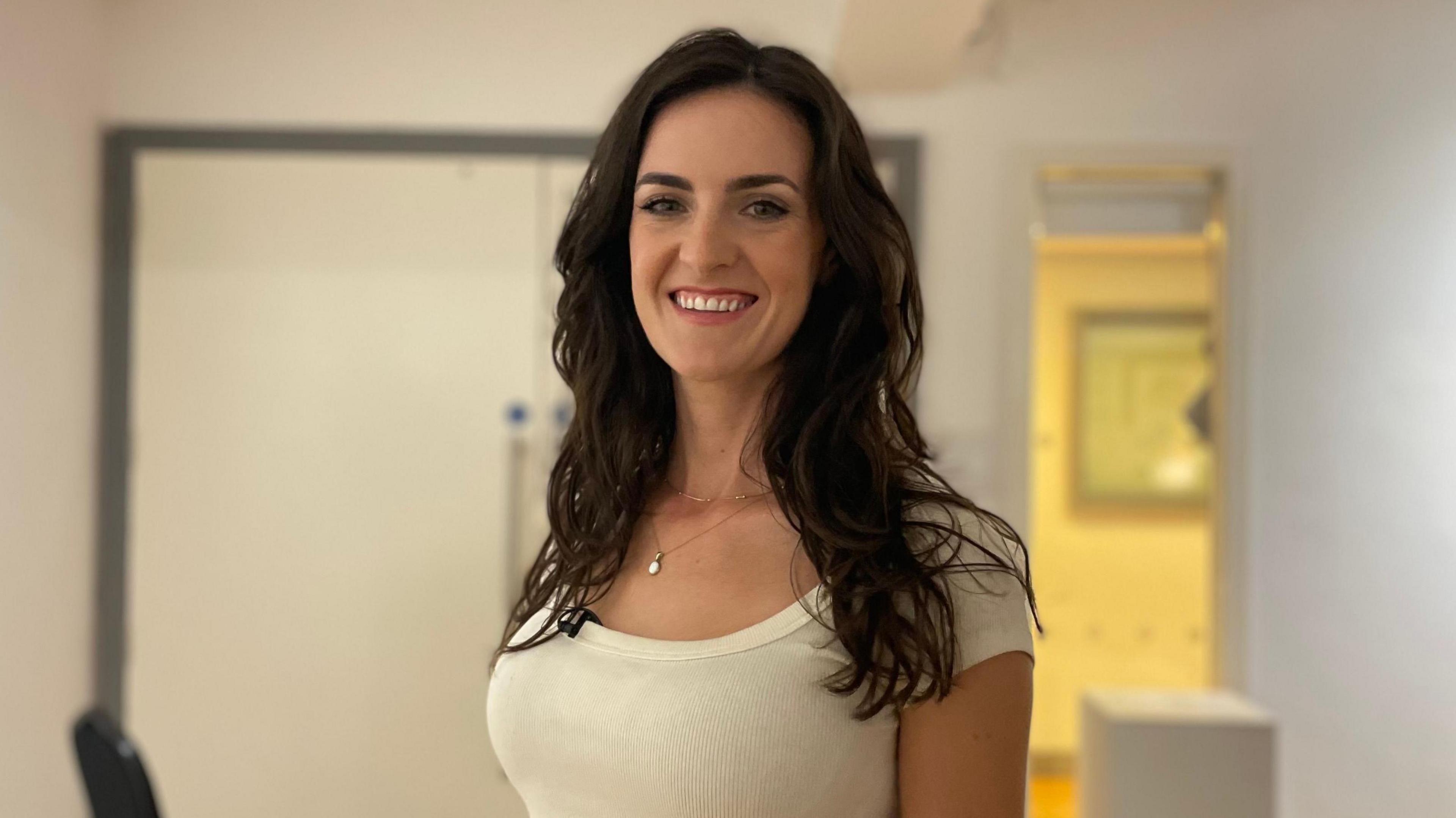 Ciara Ní É looking at the camera and smiling, with long dark brown hair. She is wearing a white T-shirt. There is a white wall and white door behind her. 
