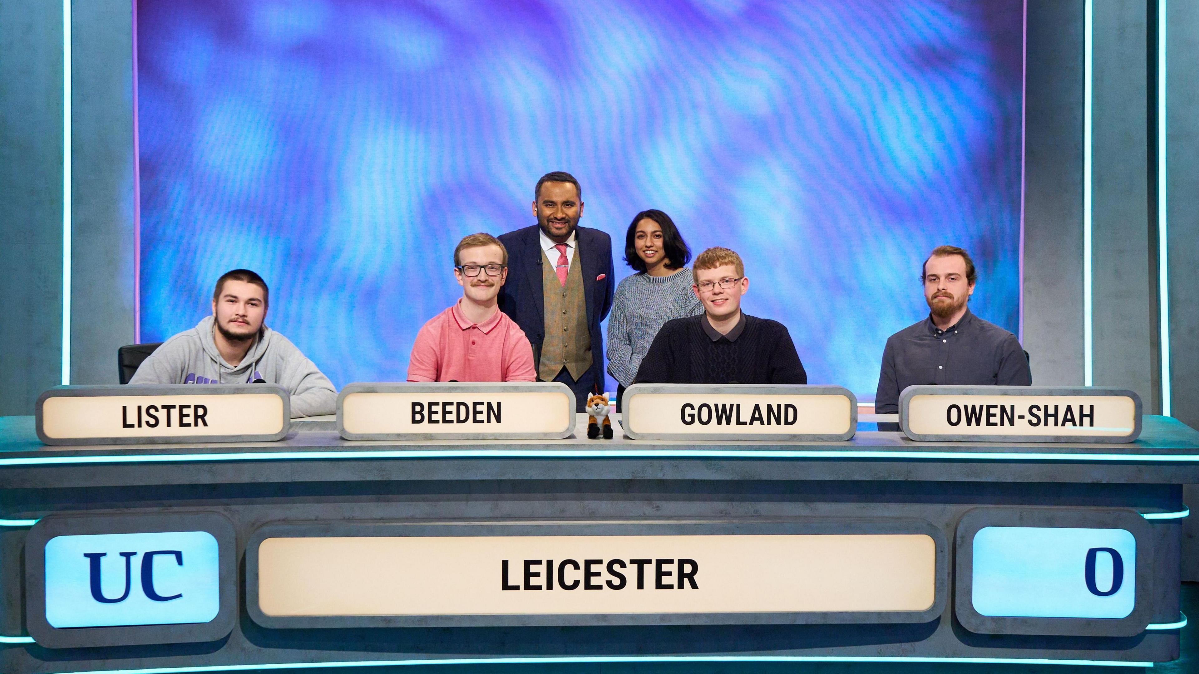 The four team members of the University Challenge team at their seats with their names displayed and zero points on the board, with presenters standing behind them to pose for the picture