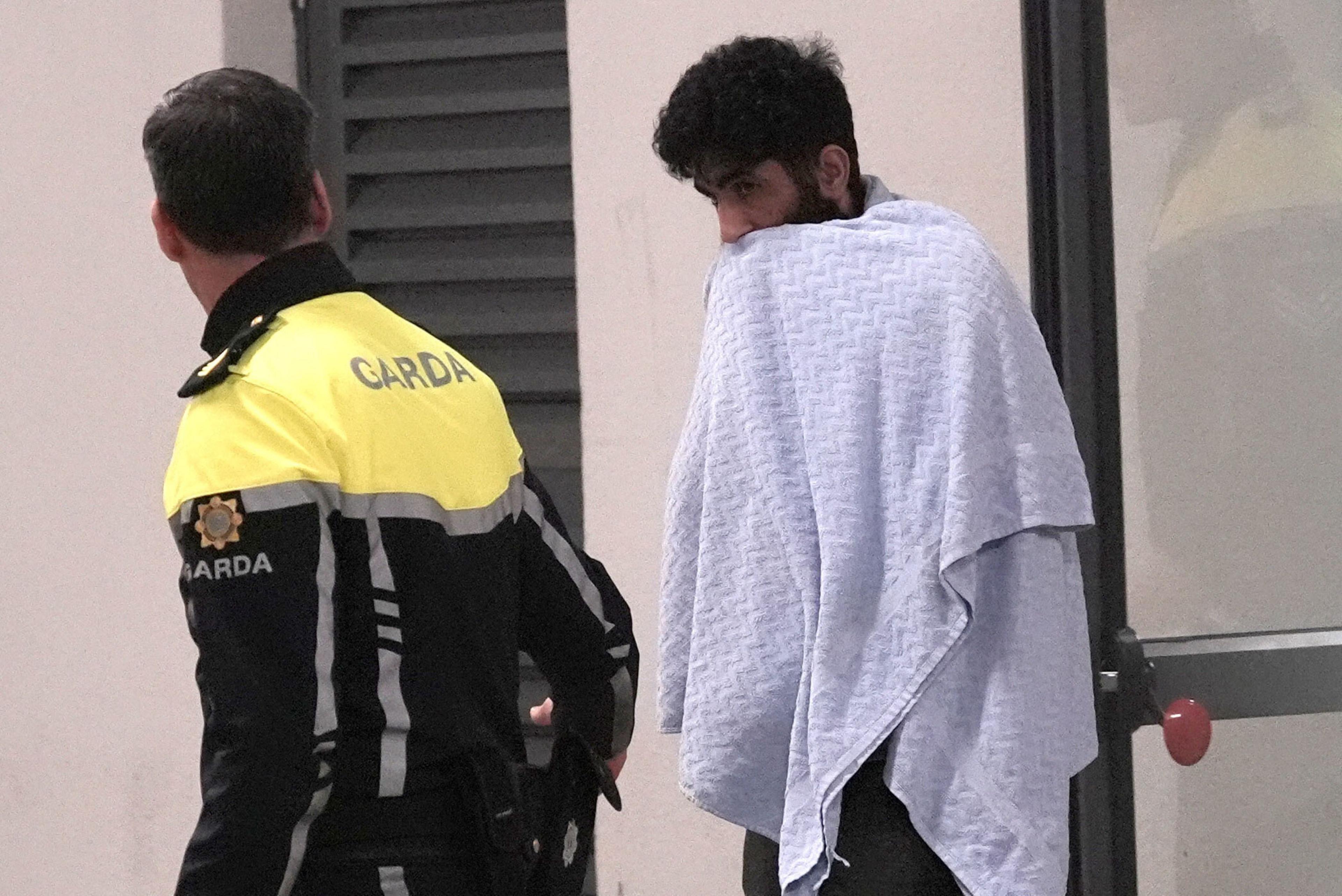 Mohammed Al Shaker Al Tamimi has black curly hair and a beard. He is wrapped in a white towel. He is accompanied by a Garda (Irish police) officer whose head is turned from the camera. He has short black hair and is wearing a yellow and black Garda uniform