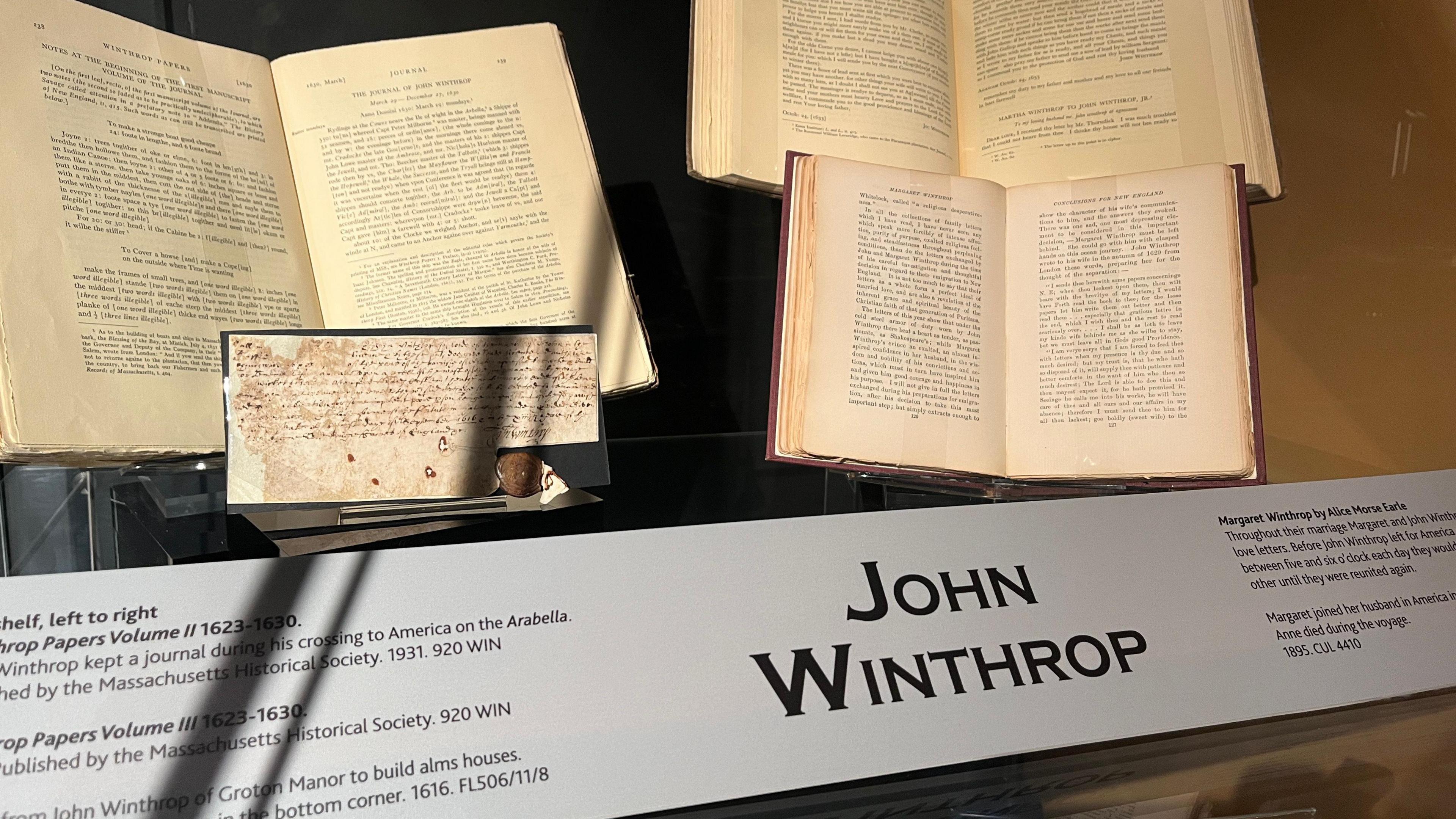 Four books are open and held in a glass display case, with a banner at the bottom reading "John Winthrop". 