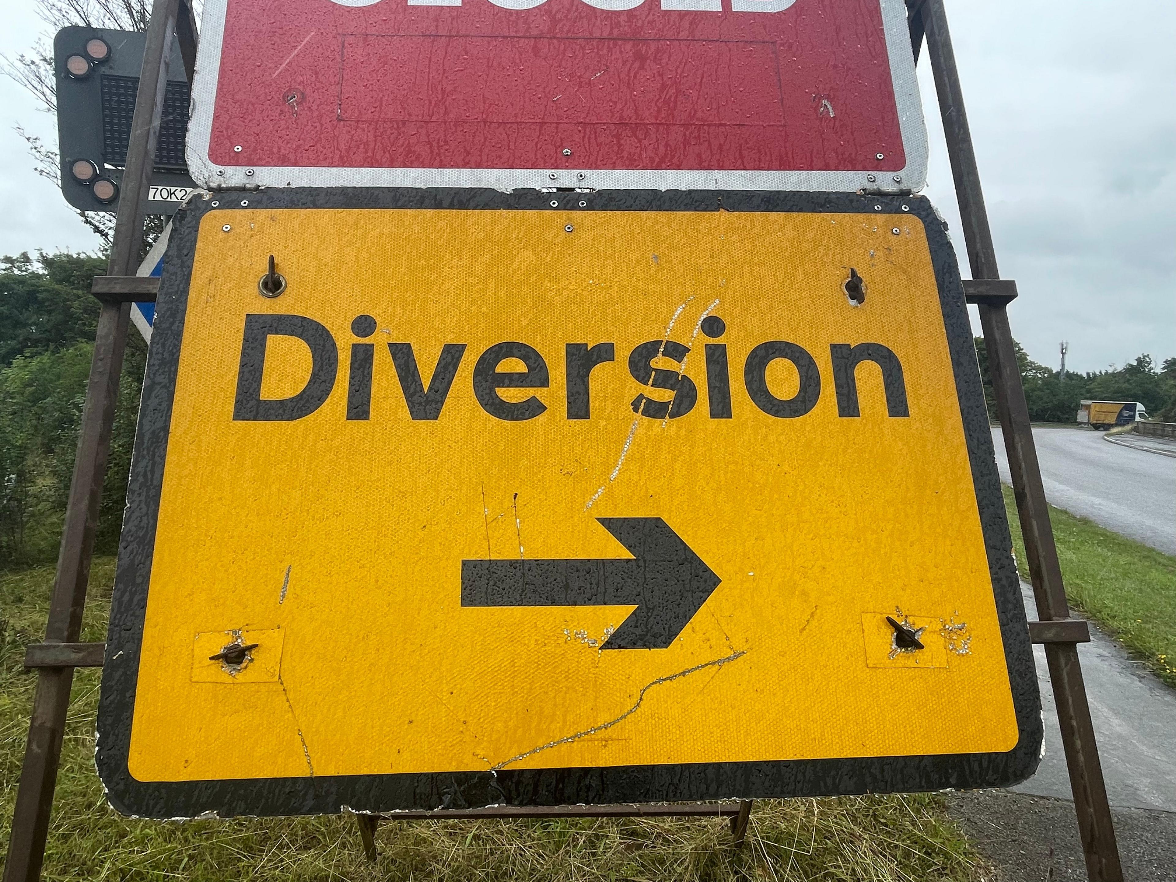 A bright yellow road sign which says "Diversion" in black with a black arrow underneath. It is resting on an verge, with a road on the right. In the far distance beyond the sign is a lorry.