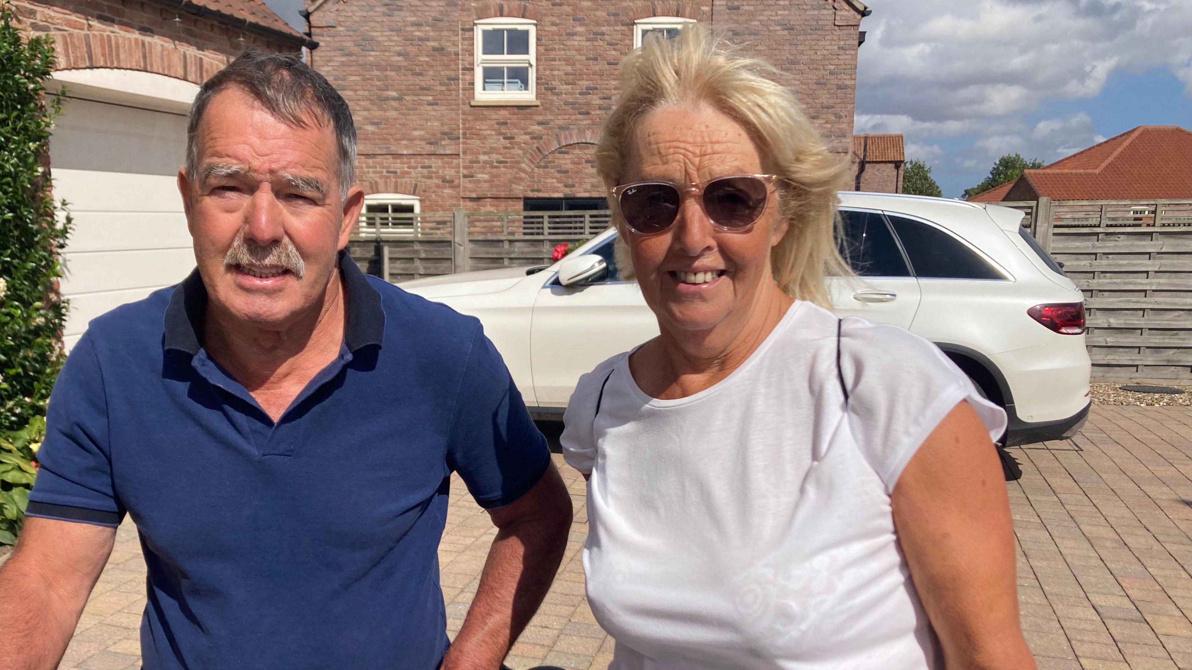 A man and a woman standing on a large brick-tiled driveway of a home, in front of a white car and a white garage door. The man has short dark hair and a grey moustache and is wearing a blue polo shirt. The woman has blonde hair and is wearing a white T-shirt and sunglasses. A large brick house is in the background.