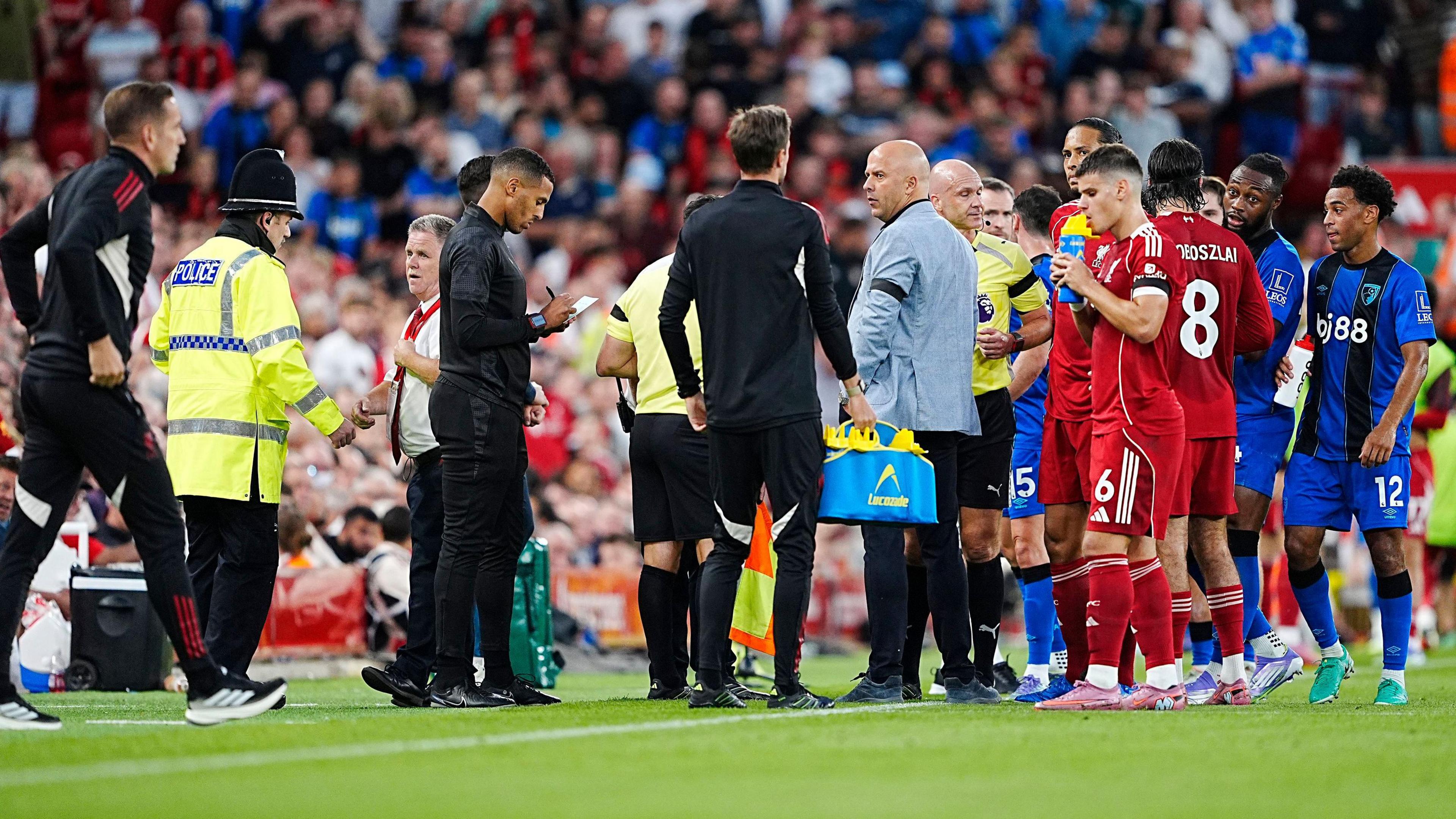 A crowd of Liverpool and Bournemouth players, including Semenyo, stand at the touchline alongside the refereeing team and Liverpool boss Arne Slot during the match pause. A policeman in a helmet and yellow jacket is speaking to a club offical while another match official writes notes on a pad and another man carries a bag of drink bottles.