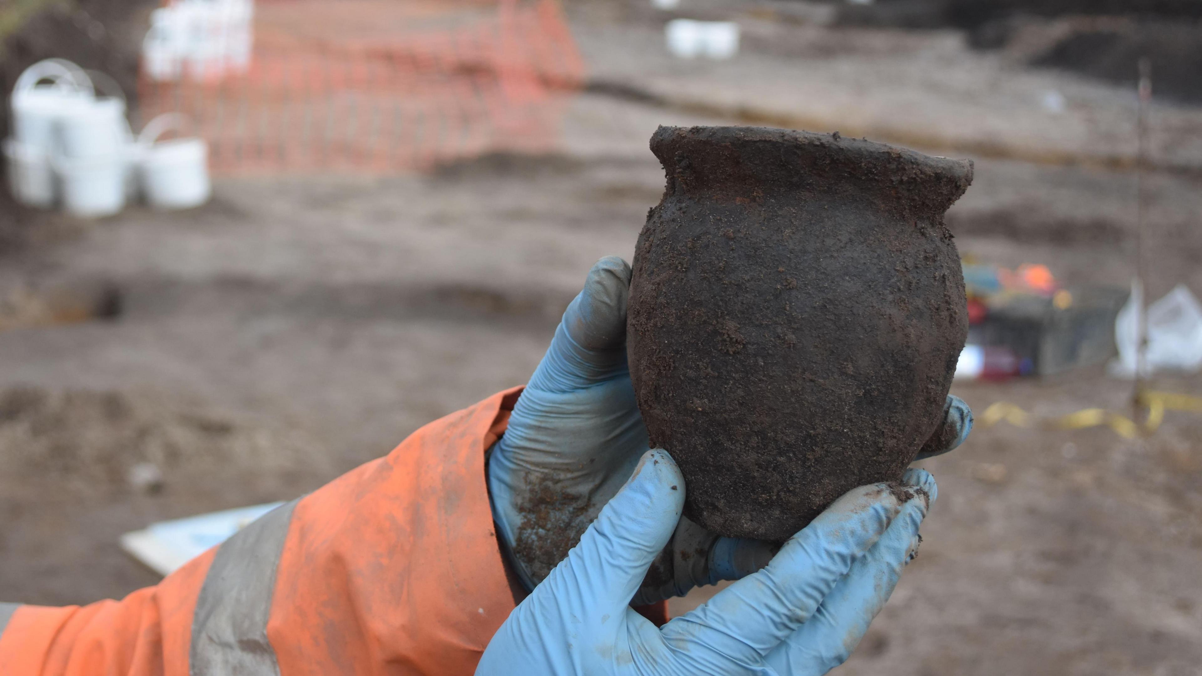 A person wearing an orange hi-vis jacket and blue disposable gloves holds up a small black pot covered in dirt.