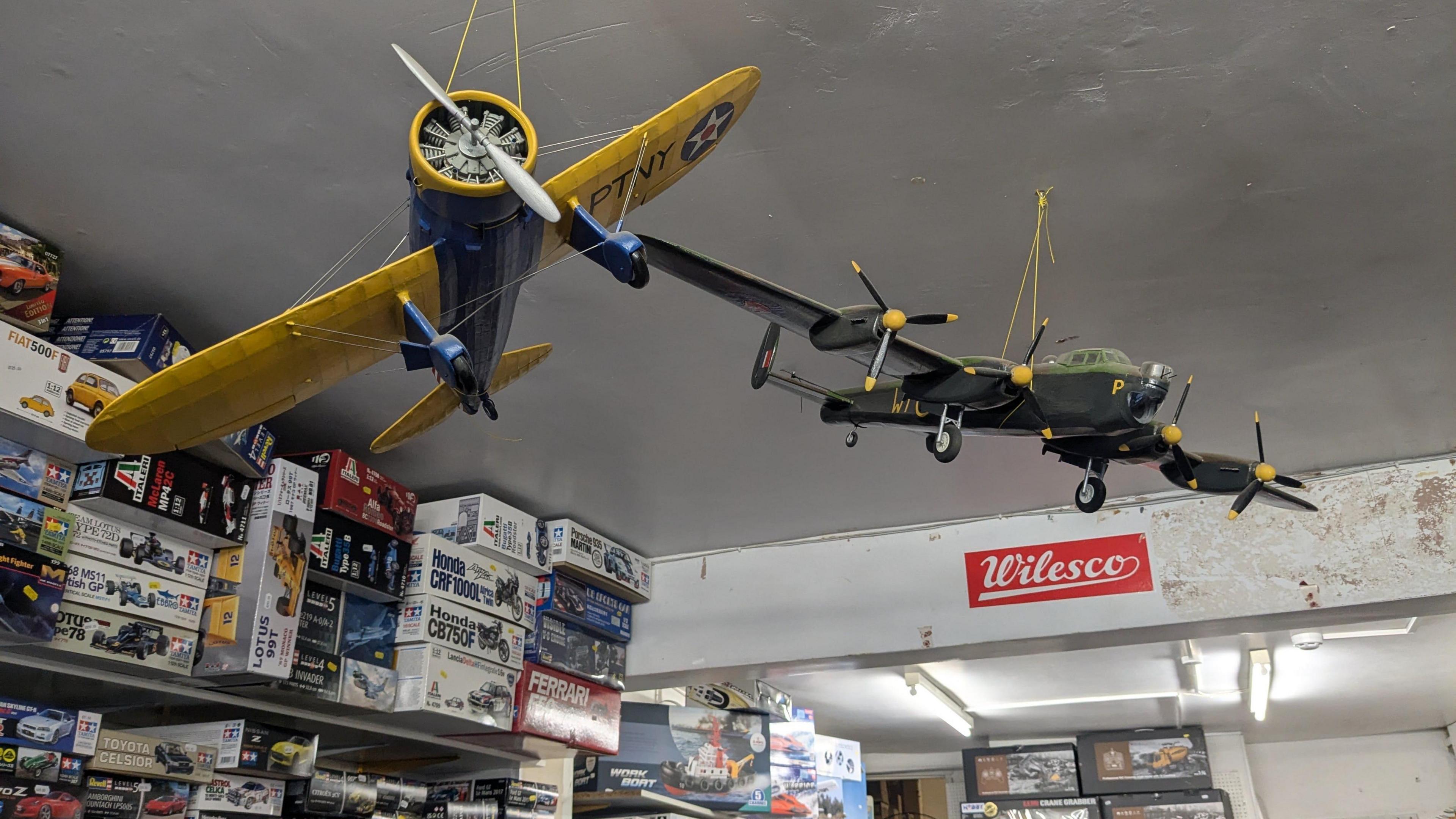 Two model aeroplanes hanging on the shop ceiling, with model making kits in the background. 