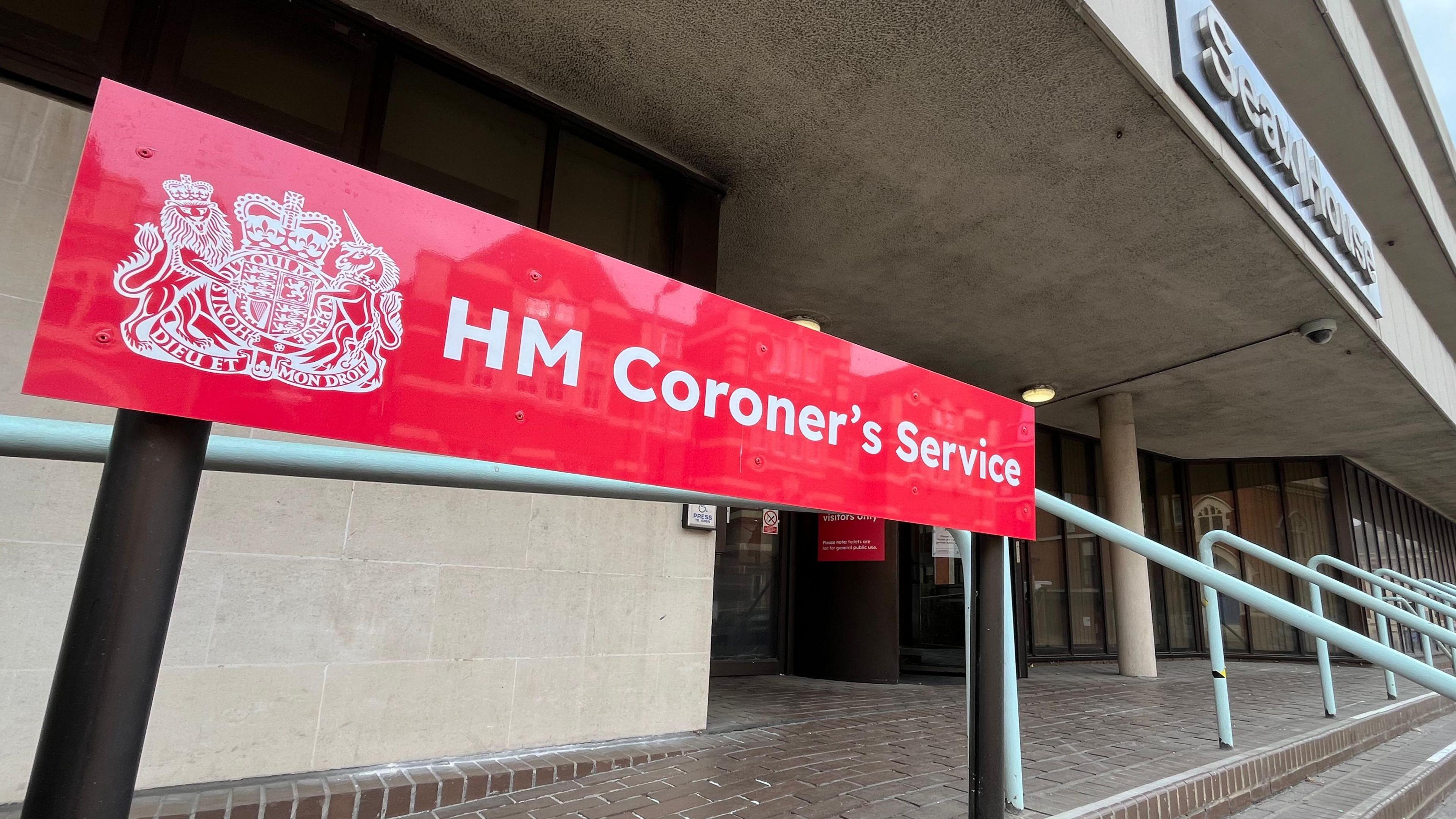 A red rectangular sign at the entrance to Essex Coroner's Court. It has "HM Coroner's Service" printed in white font. Brown brick steps lead to the entrance of the building, which is cream and has a revolving door.