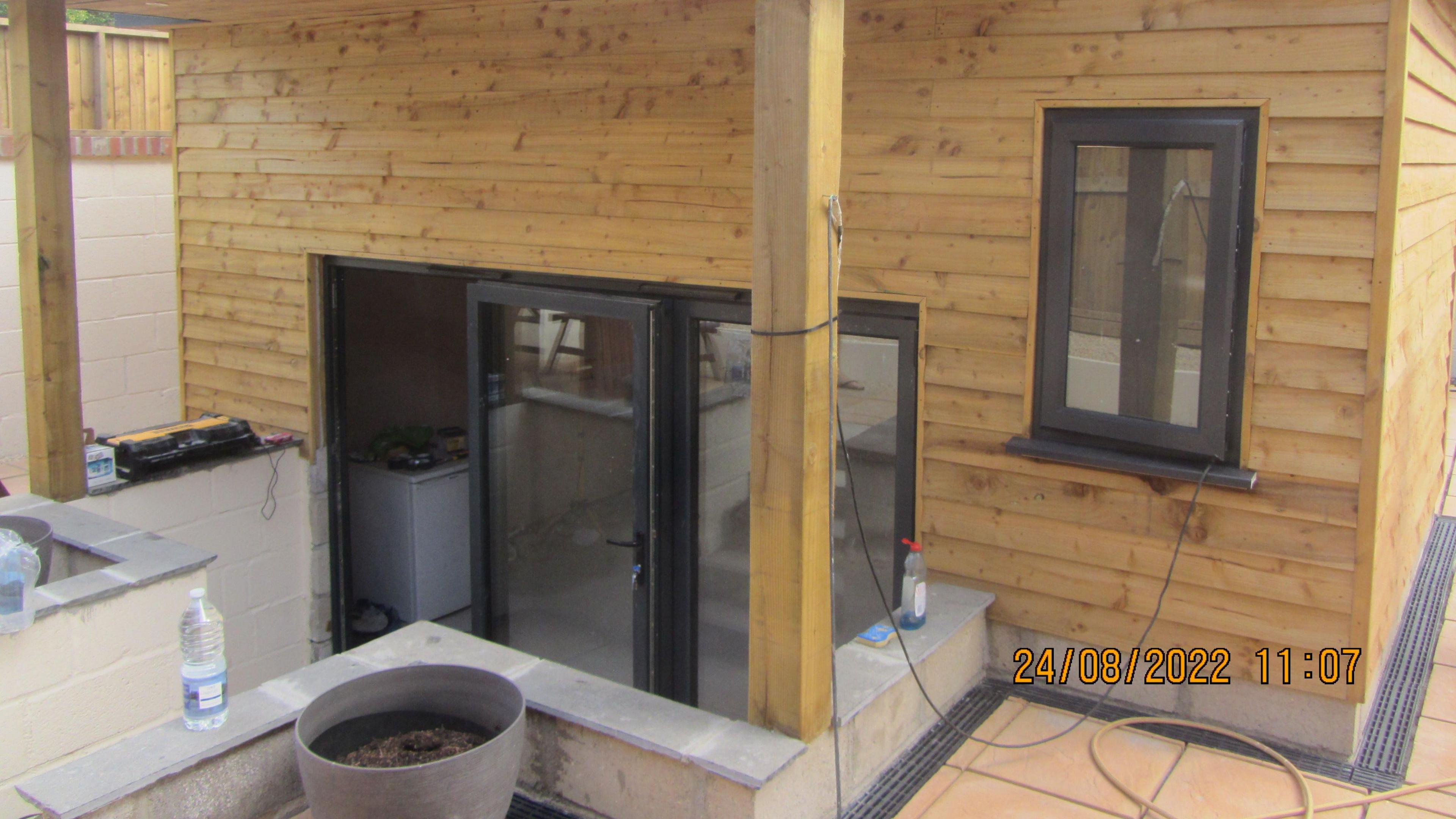 A small timber building with wooden pillars outside on a patio area. There's a flight of steps leading down towards a glass front door, and a window to one side.