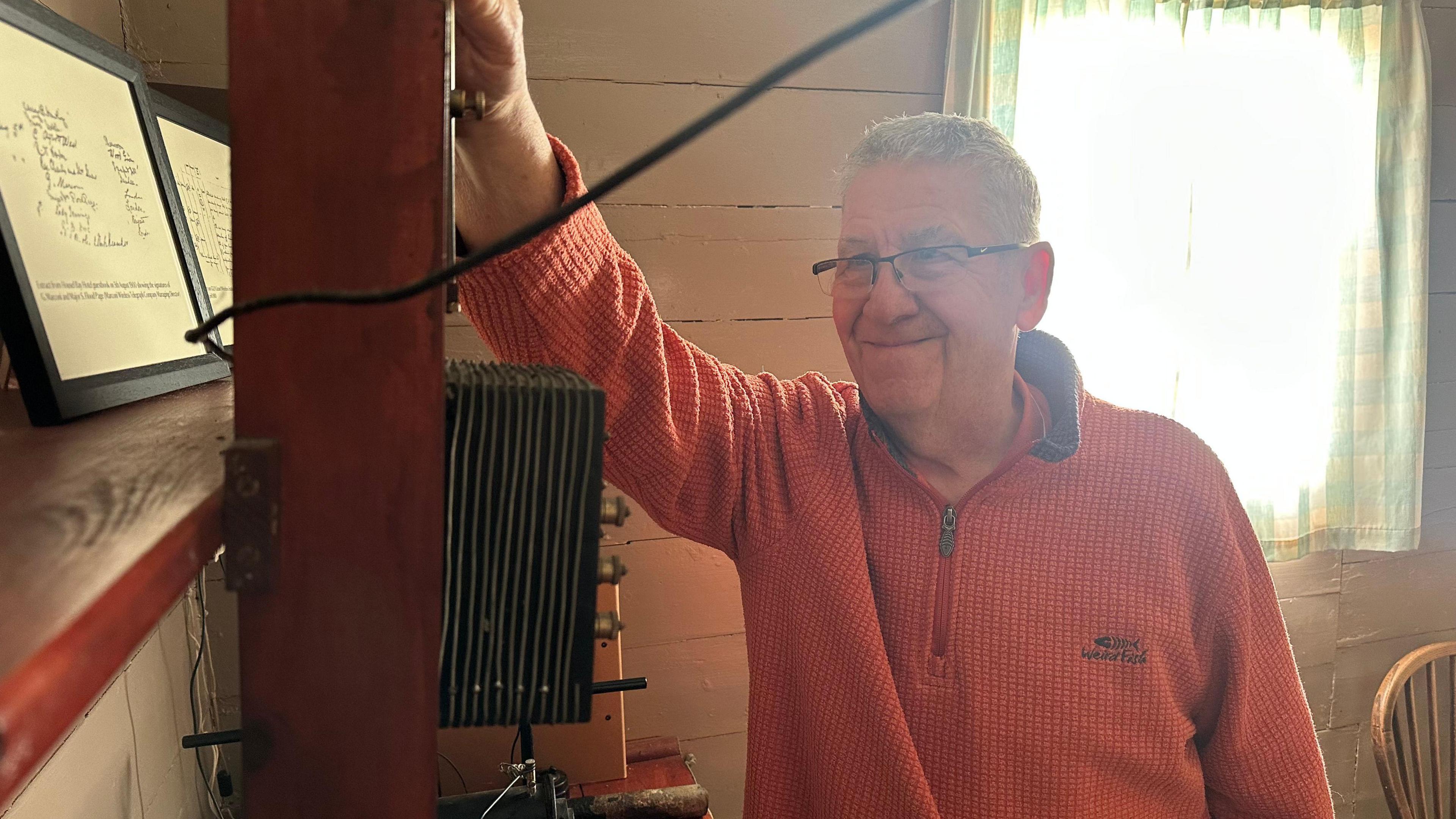 Geoff has short grey hair and is wearing a red jumper. He has his hand resting on some of the radio equipement.