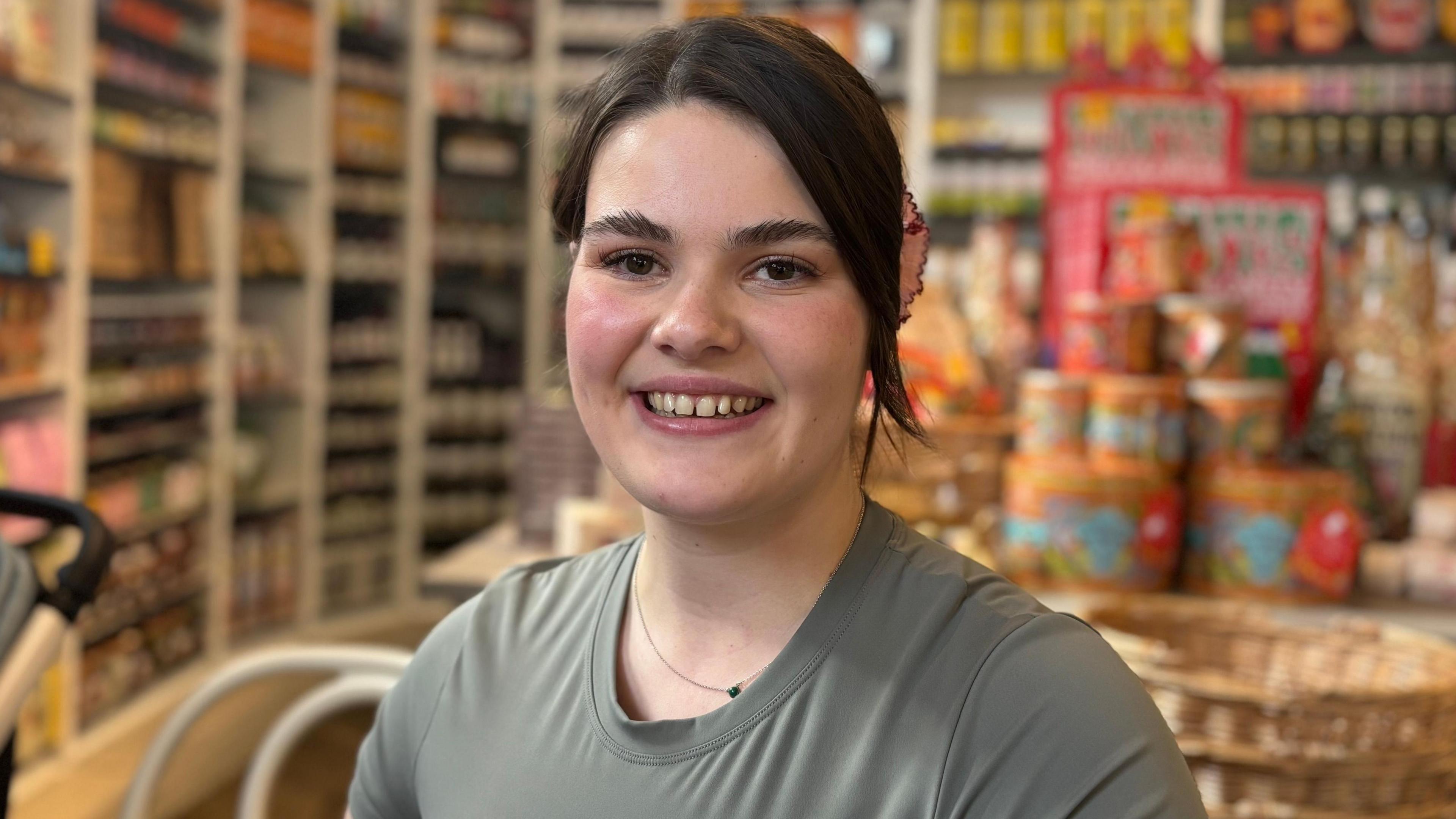 Zita with black hair tied back wearing an olive sports top. She is sitting in a cafe with food on display behind her and a chair and a pram to the left. She is smiling.