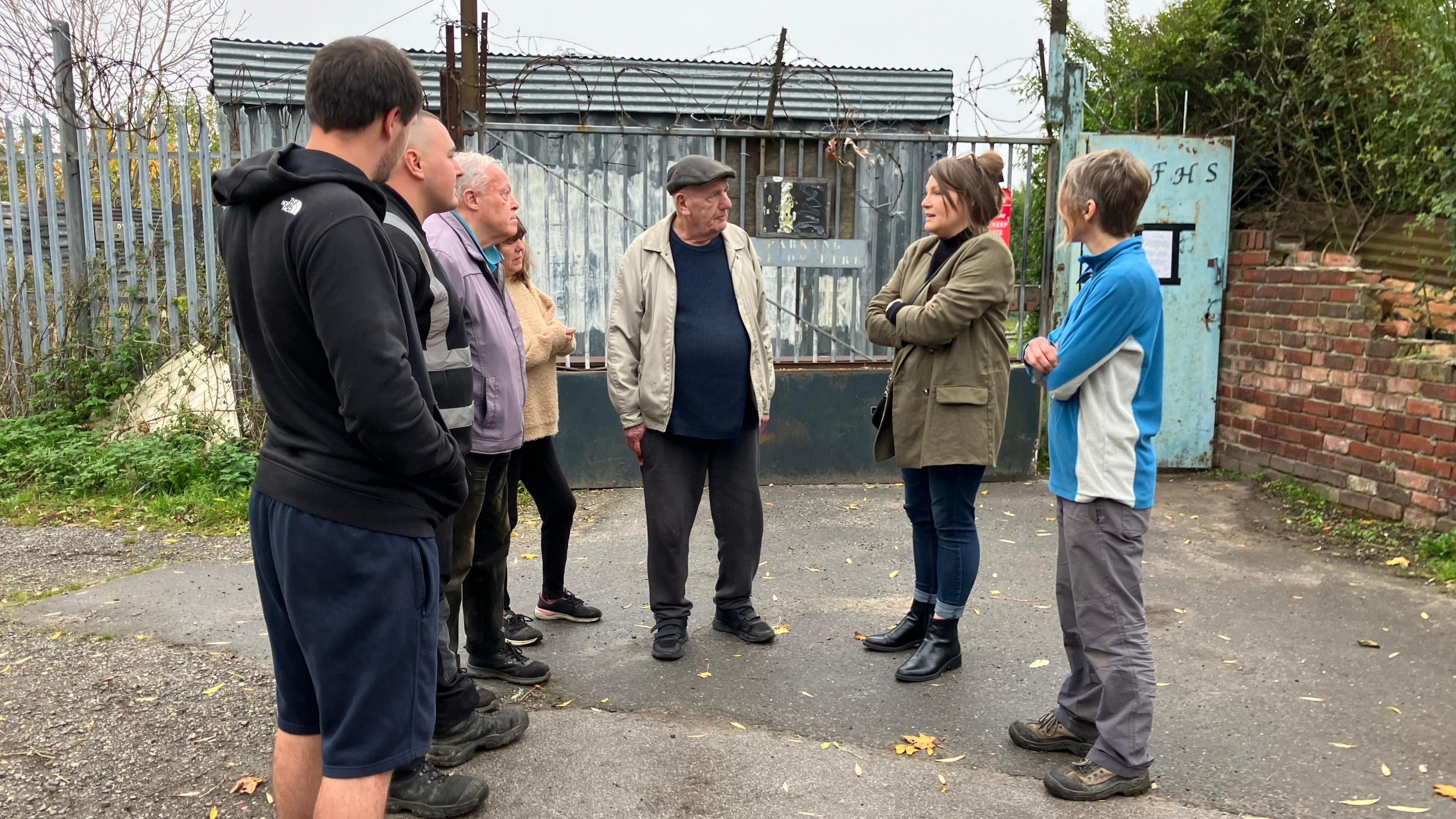 Seven plot holders stand in a semi circle in front of a metal gate with barbed wire on top.