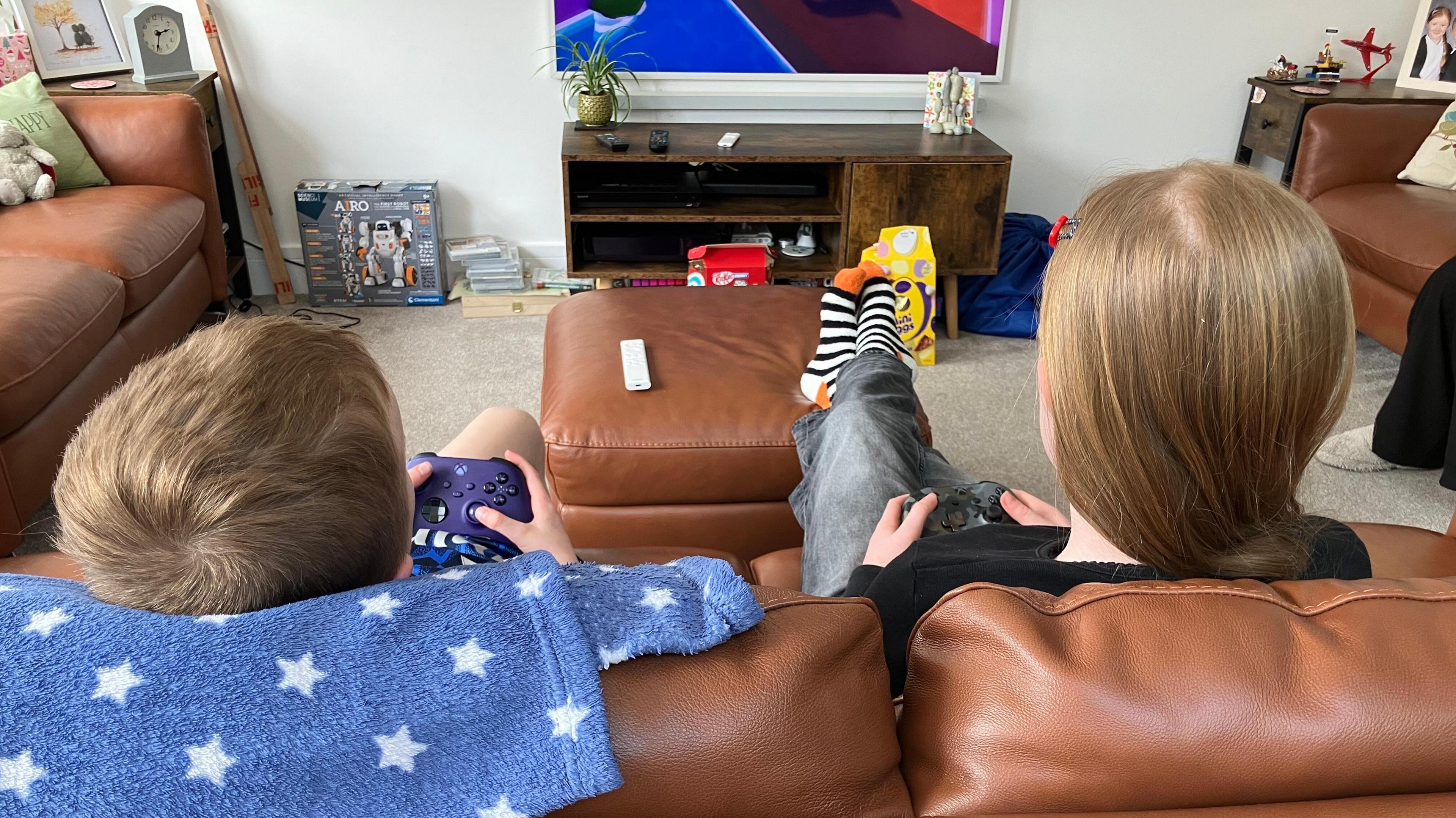 Two young children with dark blonde hair sat on a brown leather sofa playing on their Xbox, they have Xbox controllers in their hands.