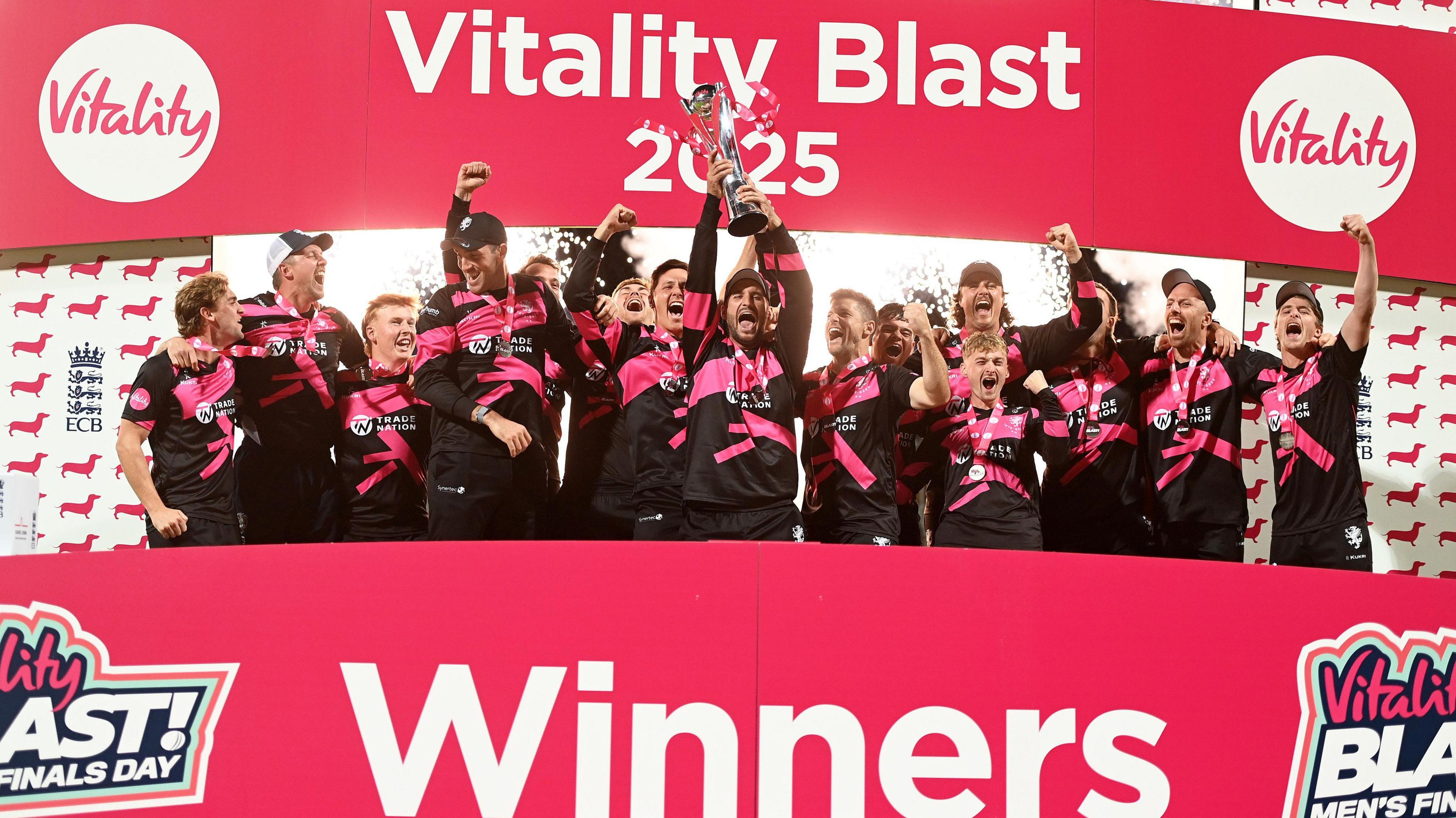 Lewis Gregory of Somerset lifts the Vitality Blast Men's Trophy, surrounded by cheering teammates, all of which are behind a red 'Winners' board.