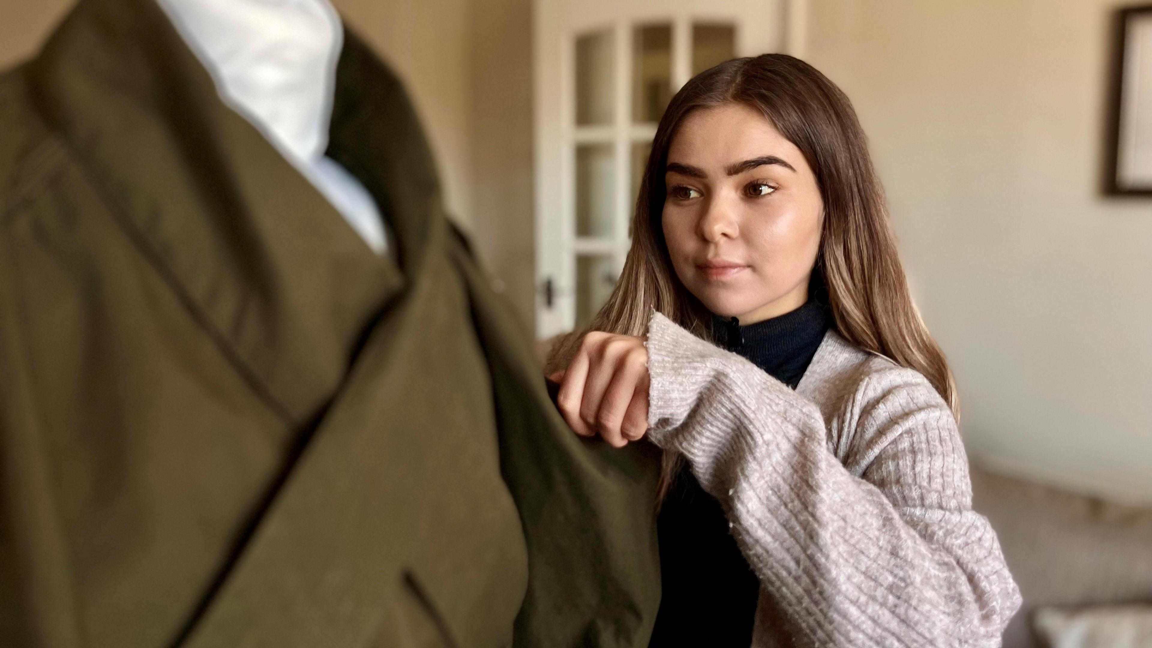 A young white woman with long brown hair and brown eyes makes adjustments to a khaki coloured coat on a mannequin 