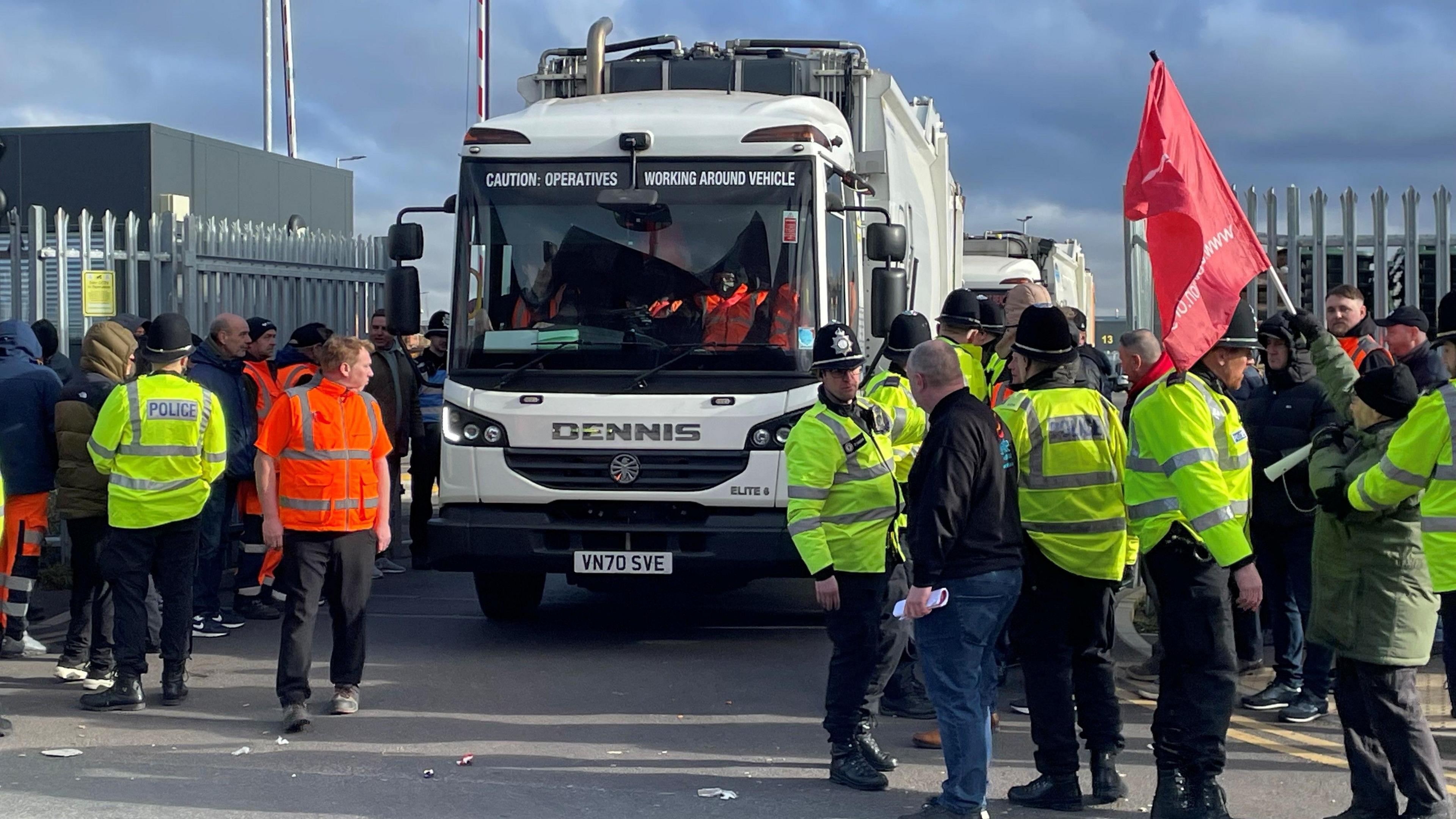 Police maintaining order at a picket line with striking bin workers in Birmingham. There are a number of people at a set of gates and a DENNIS bin lorry coming out as workers and police look on. 