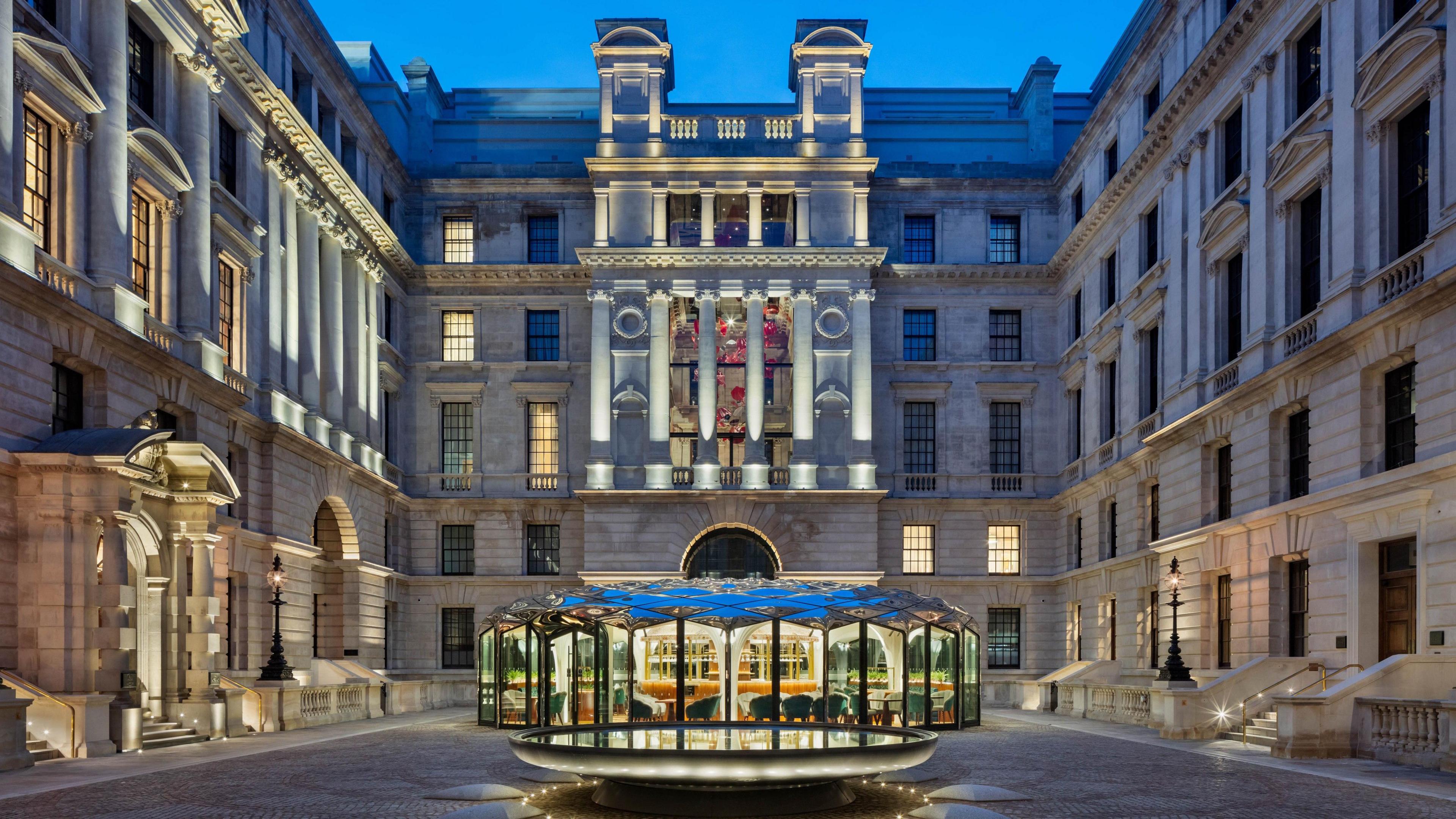 The exterior of the building is lit up at night with rows of columns and a glass building on the ground floor that contrasts with the brick building surrounding it. 