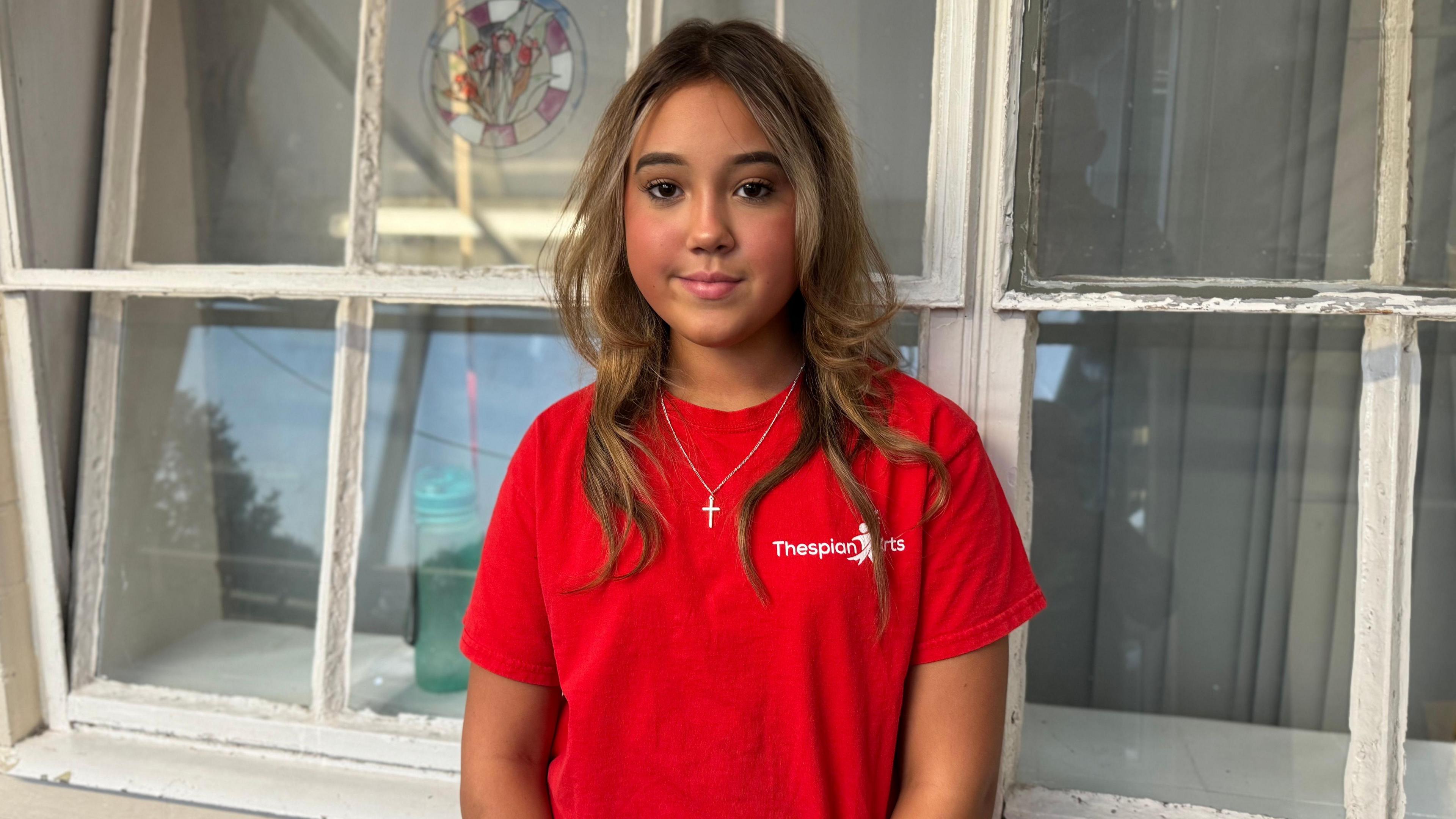 Isabelle looking at the camera. She is wearing a red T-shirt and is standing in front of a window. 