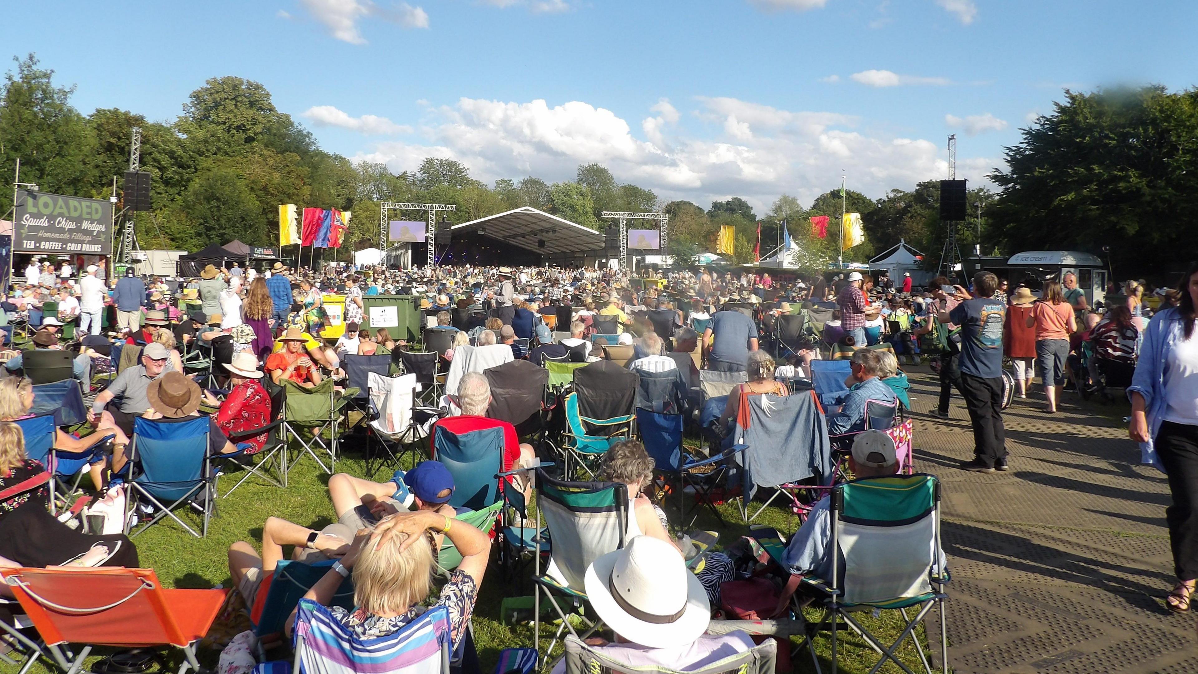 People sitting in chairs at the Cambridge Folk Festival with the main stage in the background