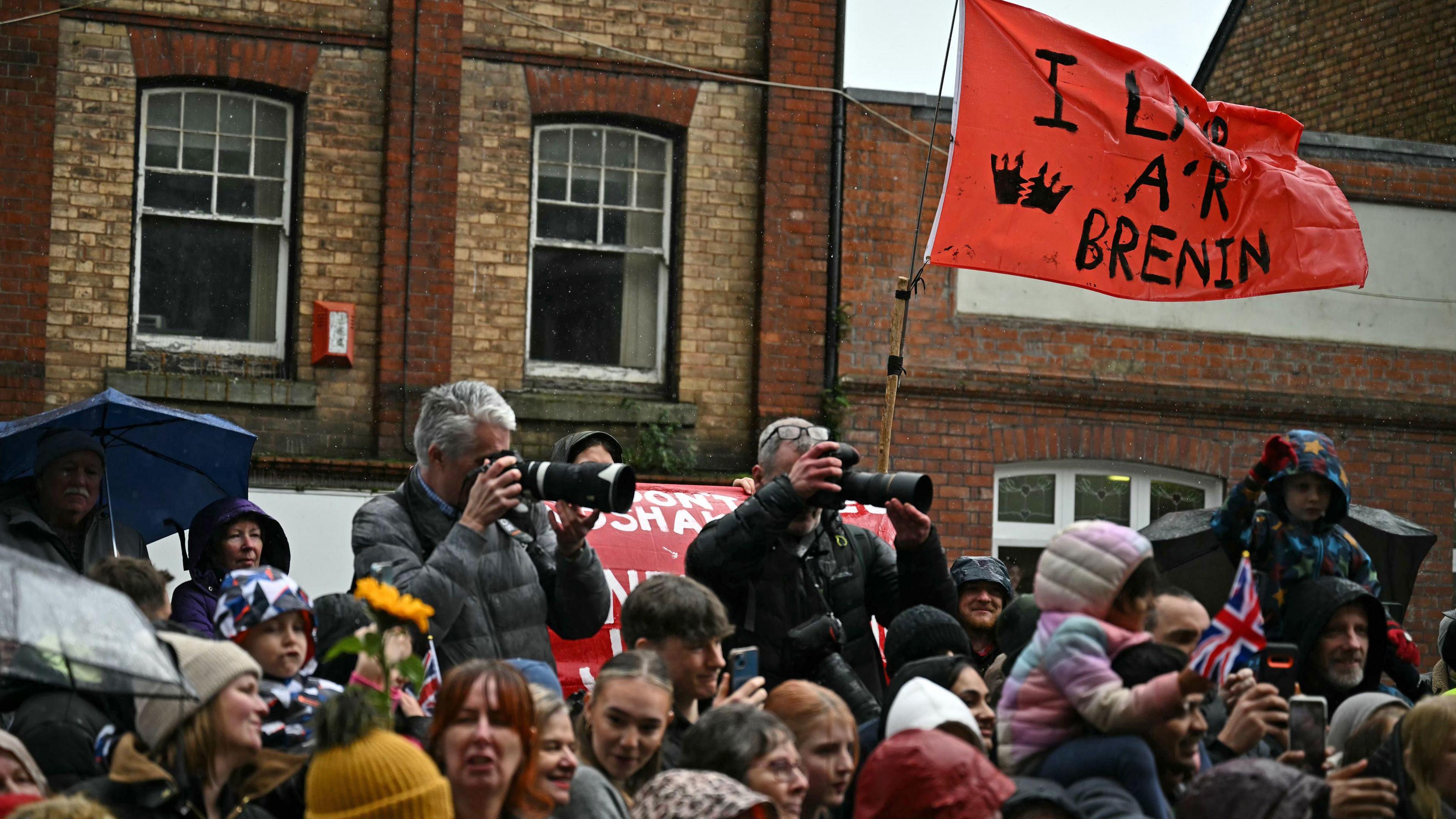 An anti-monarchy demonstrator waves a flag as the Prince and Princess of Wales greet well wishers during a visit to Pontypridd Market. There are dozens of people watching and waiting to meet the royal couple, the anti-monarchy flag is being waved from someone near the back of the crowd. It says ‘I lawr â’r Brenin’ translated as down with the king.