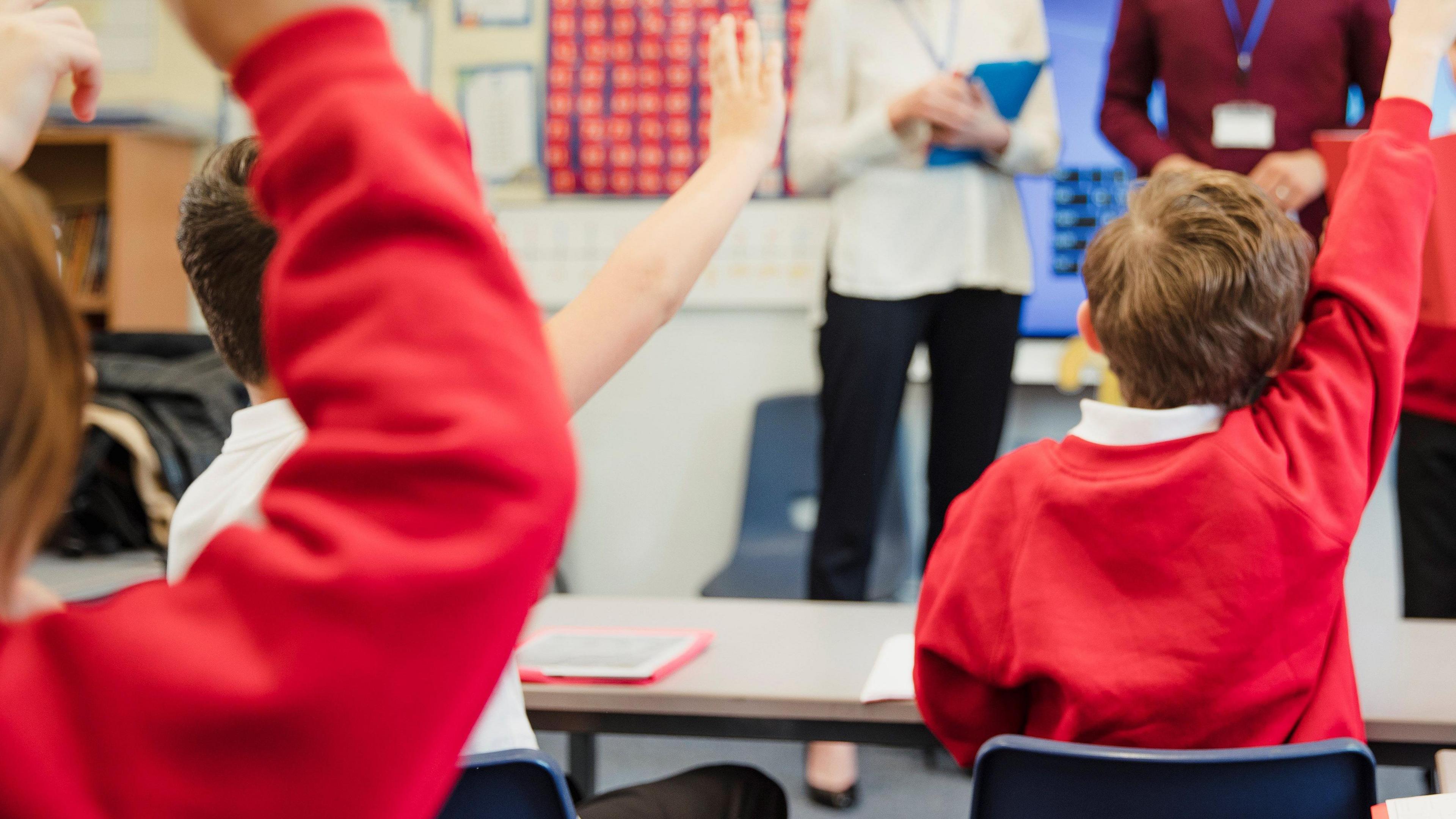 An elementary schoolboy wears a school uniform and presents his work to the class with the support of his teachers beside him.