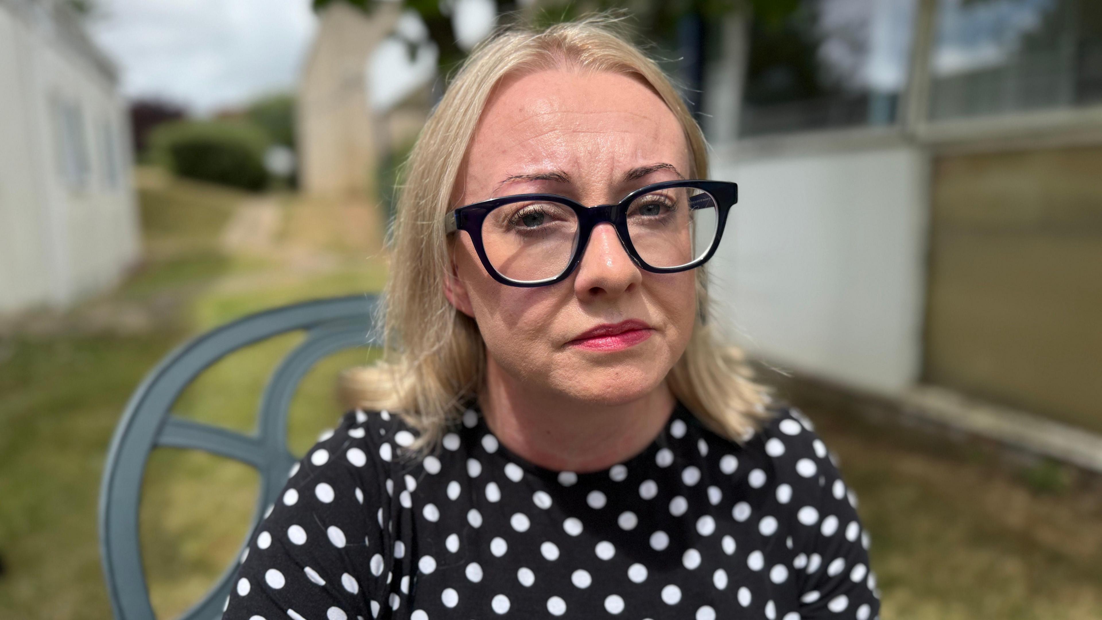 Julie Hogg looking into the camera while sitting outside hospital buildings. She has cropped blond hair, thick rimmed black glasses and is wearing a black top with white polka dots.