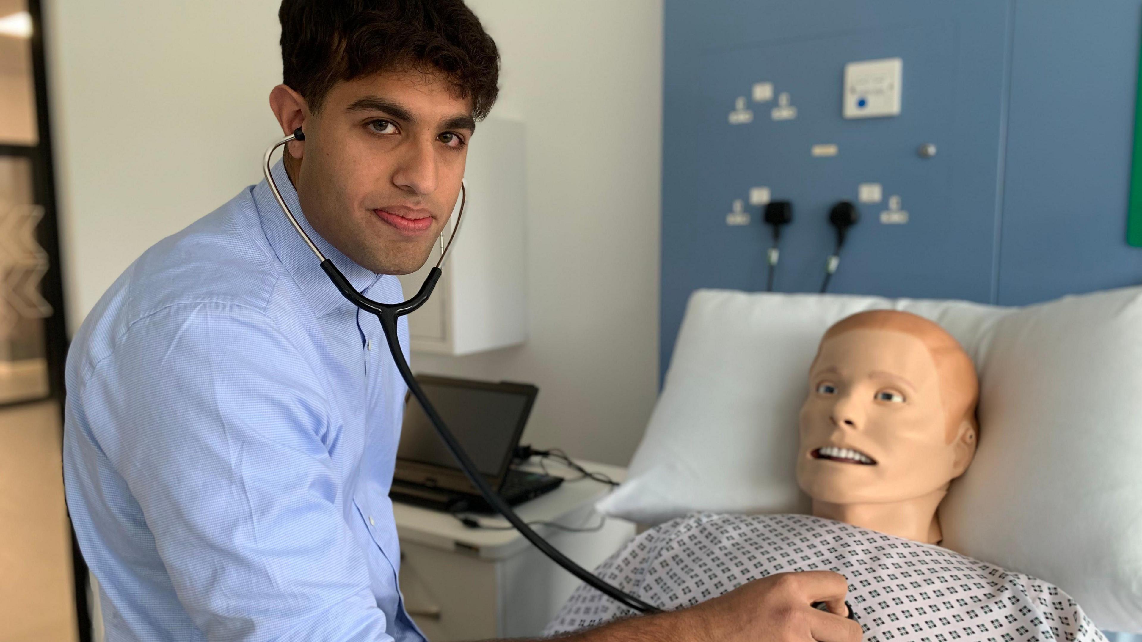 A young man with dark hair and a blue shirt puts a stethoscope to the chest of a model of a patient used to train young doctors in a hospital bed.