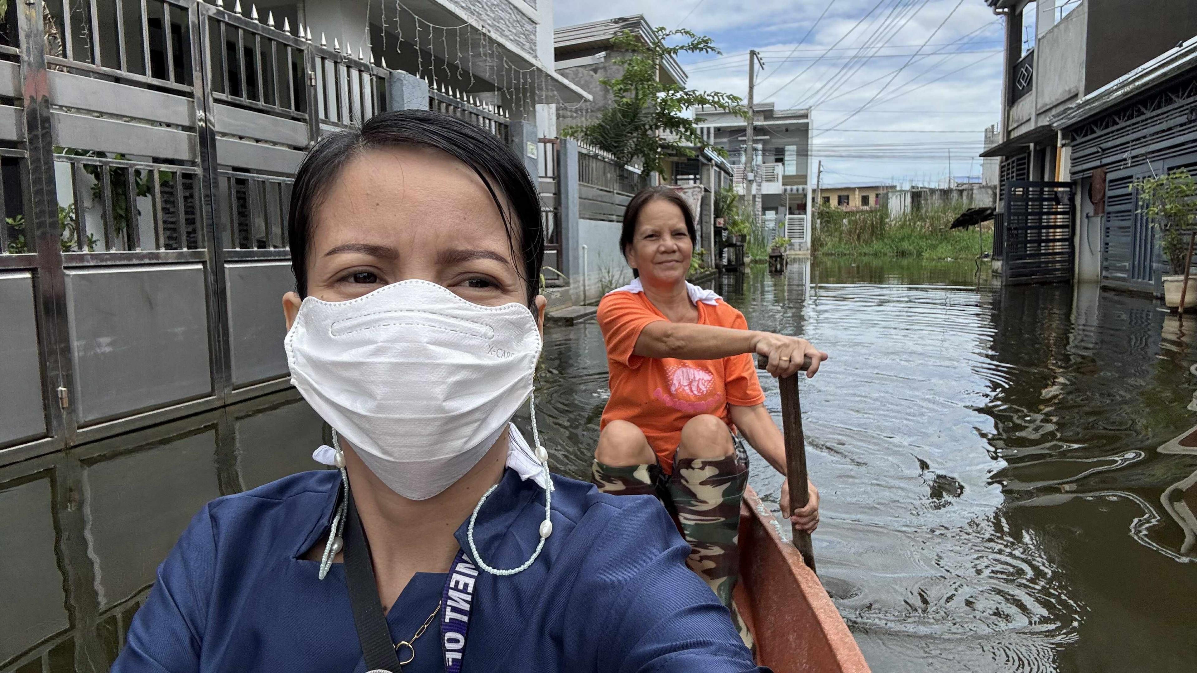 Woman wearing face mask riding a boat through a flooded street with another woman paddling behind her
