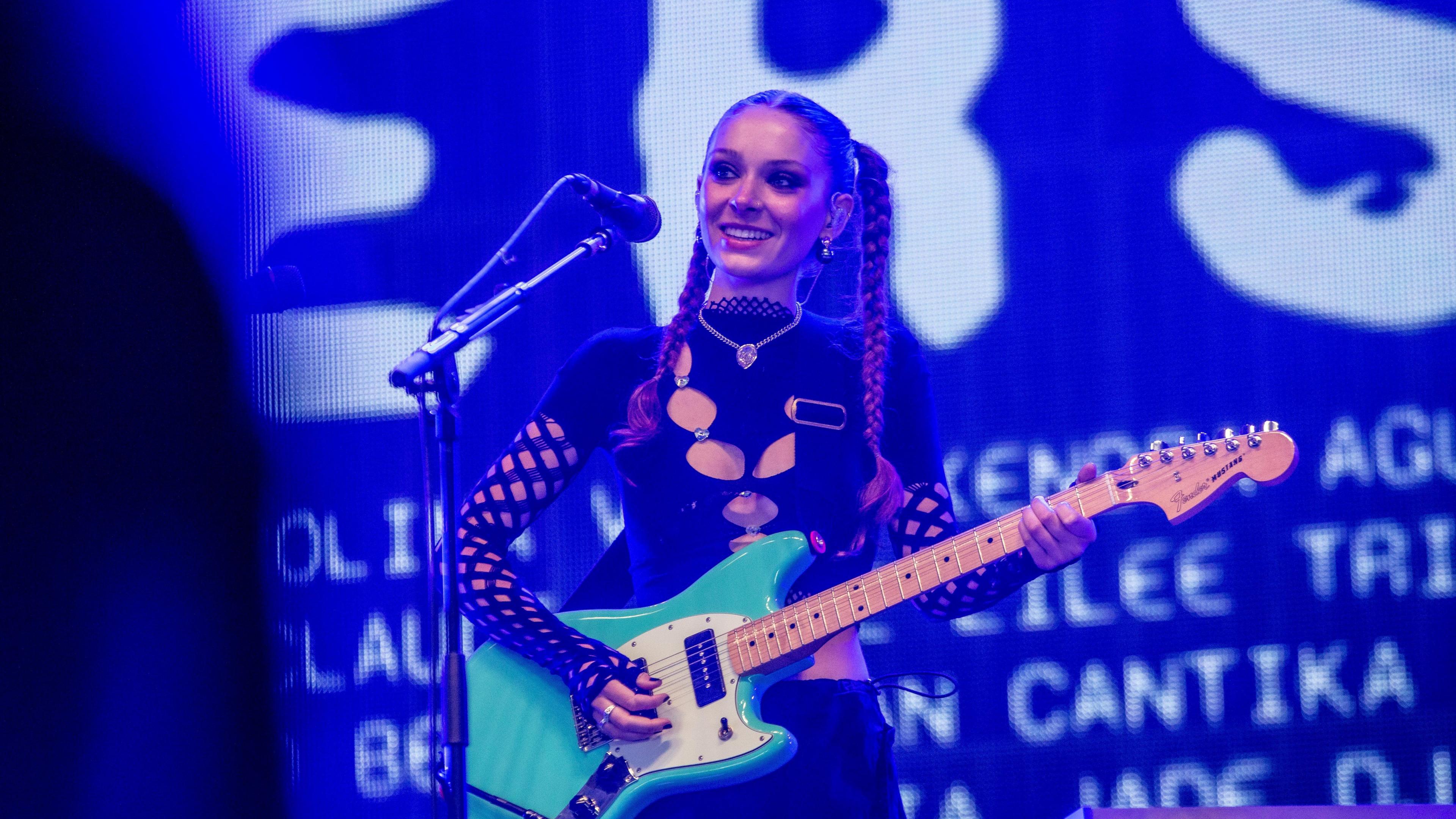 Holly Humberstone performs on stage. She is dressed in all-black and playing a guitar. She is bathed in blue light and the backdrop has white writing on it.