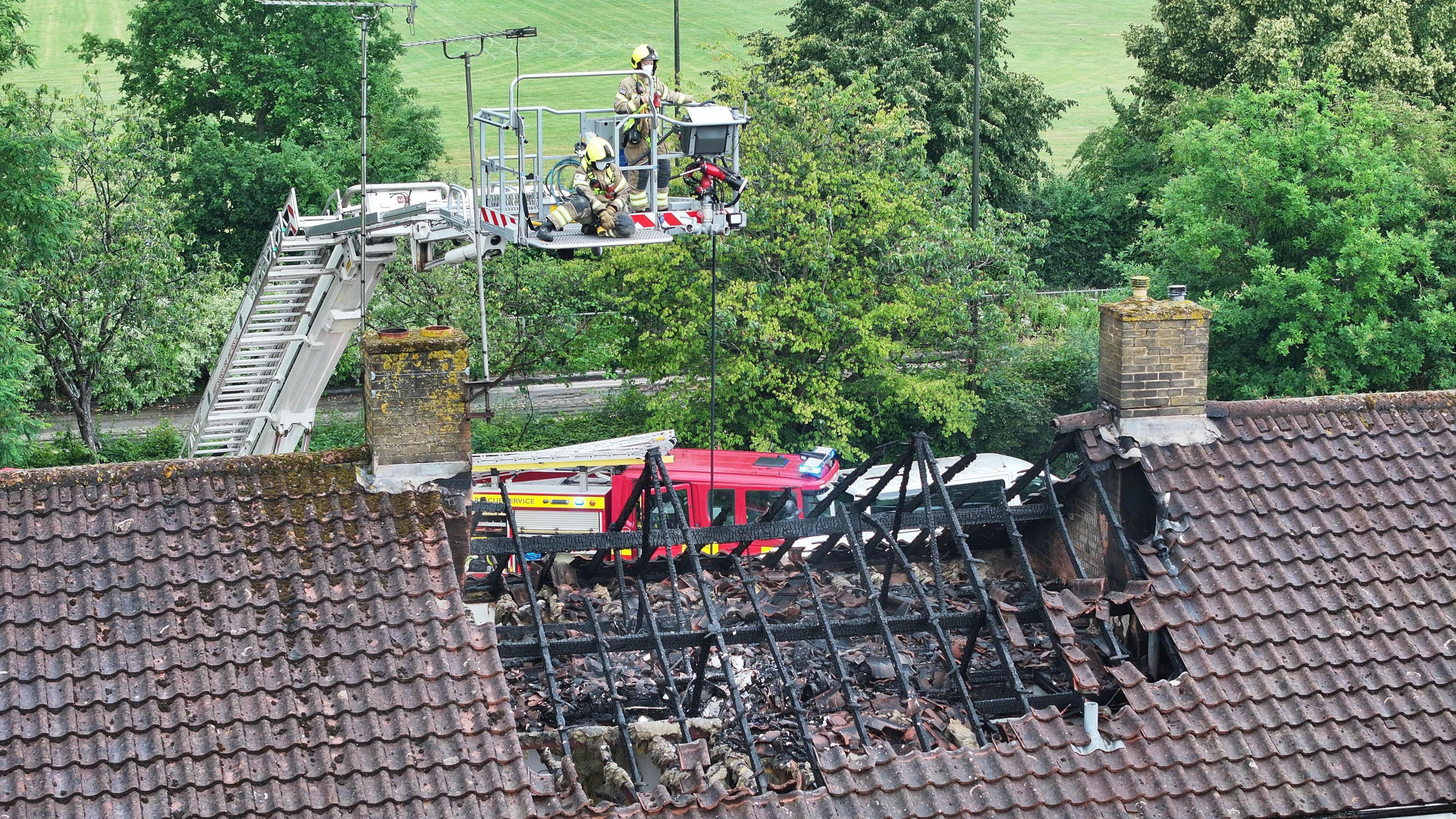A fire crew on an aerial ladder looking over a damaged section of the roof of a house. The timbers in the roof are badly charred, and there is rubble in the hole caused by the damage.