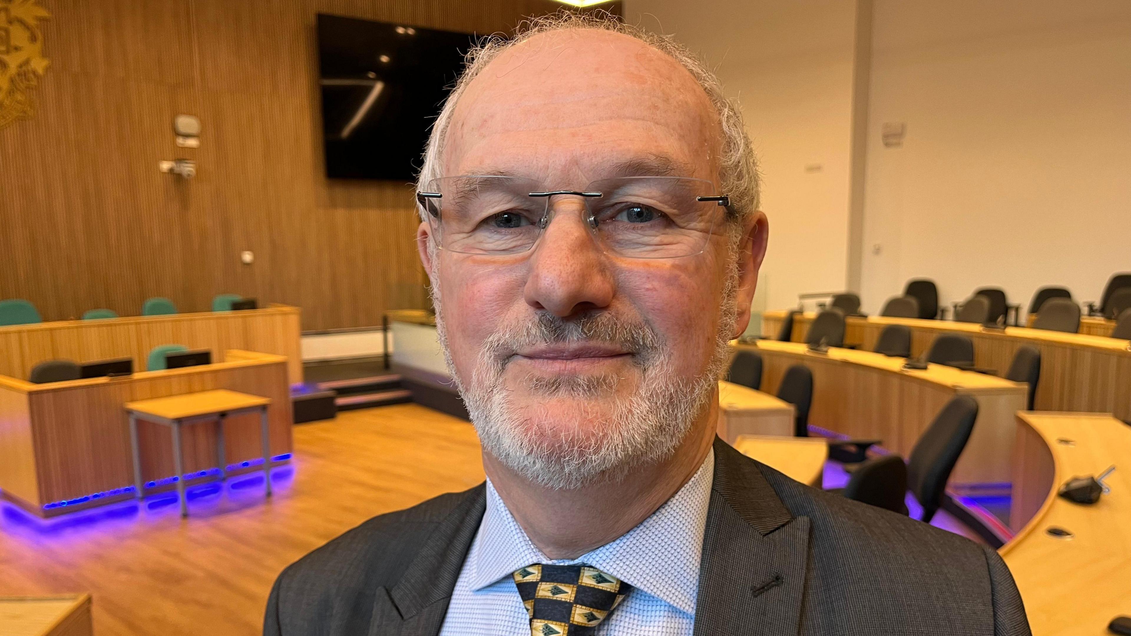 Stephen Davies smiles at the camera as he stands in the council chamber. He is an older man, with short grey hair and a short beard, and is wearing glasses and a suit. The room is furnished with light wood desks at the front of a room, surrounded by a semi circle of light wood desks. There is purple underlighting beneath the desks. 