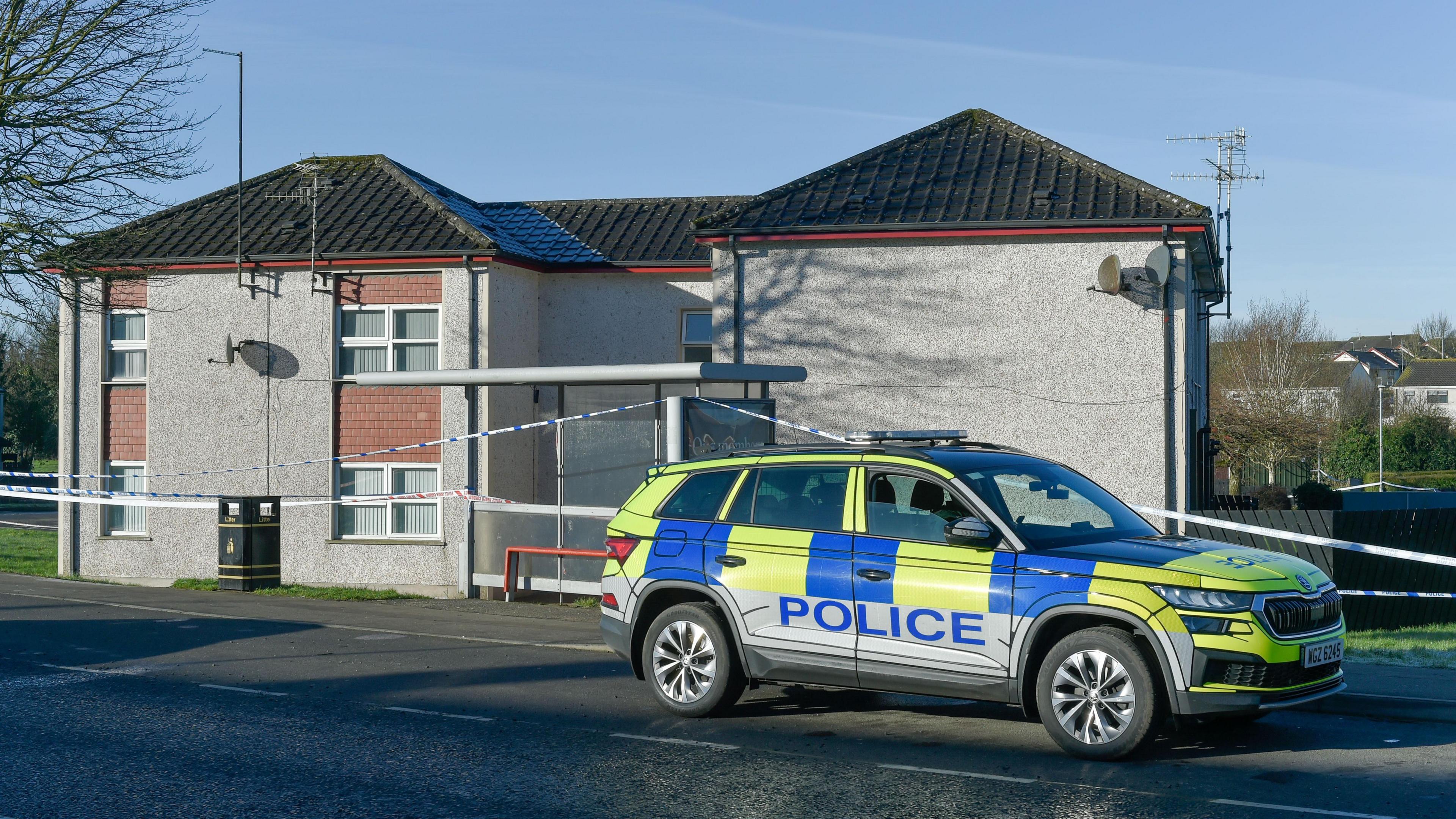 A police car parked on a road in front of a property. There is police tape in front of the property and a blue sky in the background.