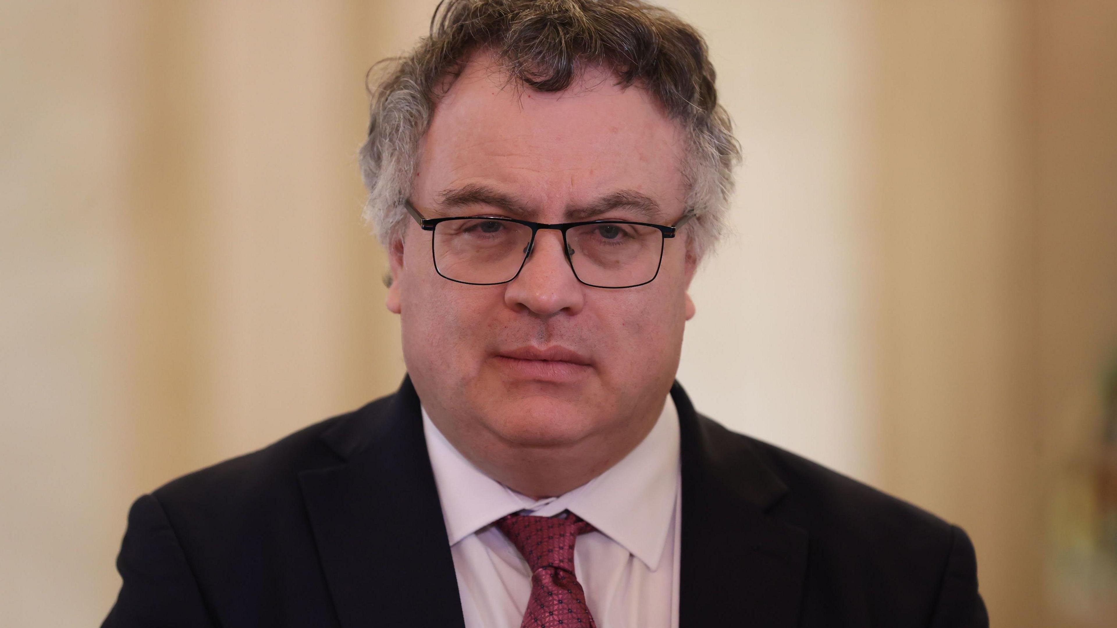 Man with curly grey hair and glasses, wearing a pint shirt, maroon tie and dark jacket.