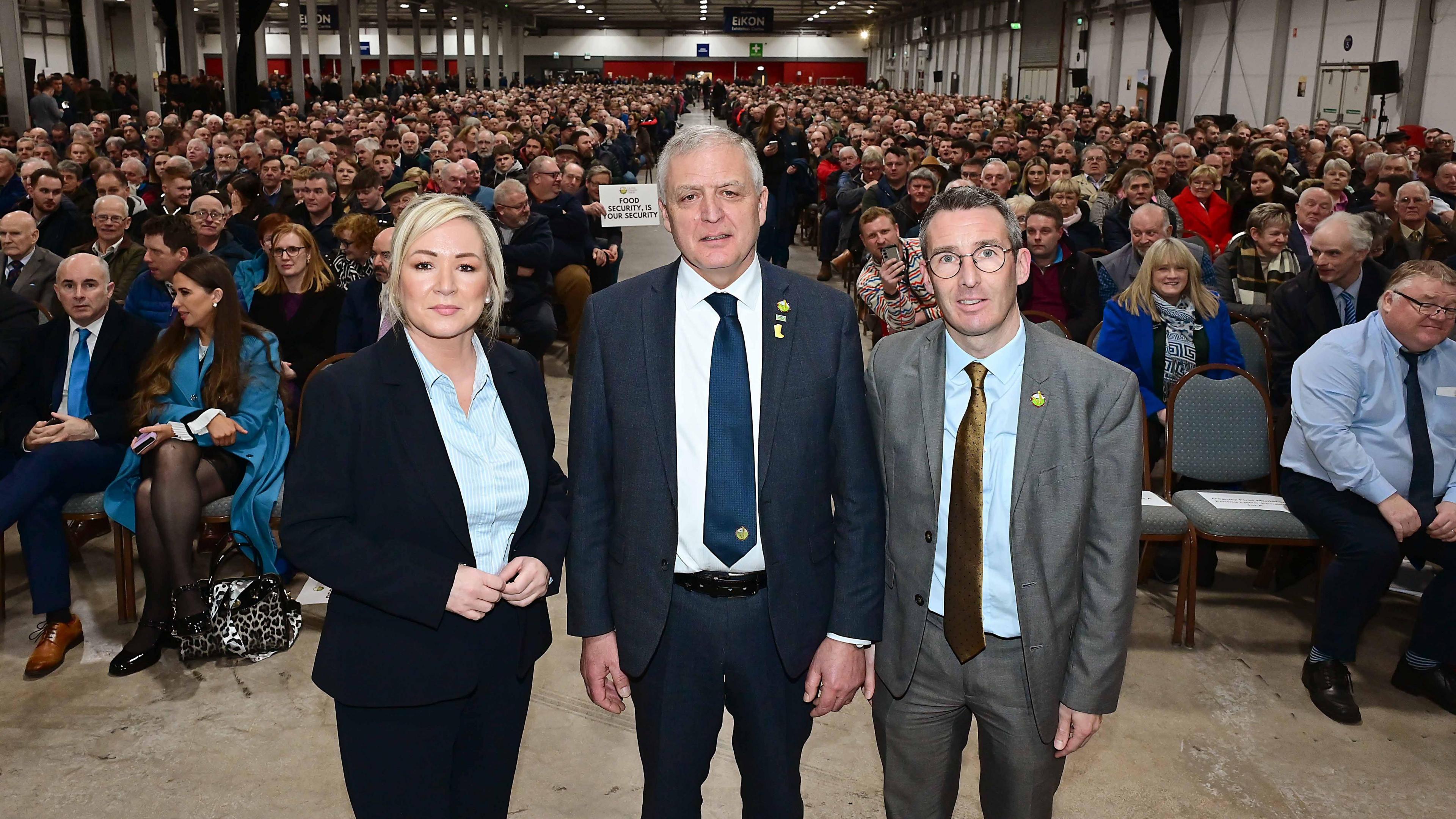 First Minister Michelle O'Neill, dressed in a navy trouser suit and blue shirt, on the left, with Agriculture Minister Andrew Muir, dressed in grey suit and brown tie, on the right. In the middle is William Irvine in a navy suit and navy tie. He has grey hair. The exhibition is full of people sitting on chairs.